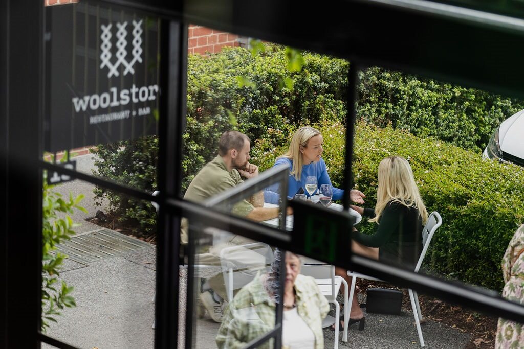 View through black T-bar windows, looking out to three young people sitting around a small white table, behind them are some lush green bushes. A black sign above states "Woolstores Restaurant & Bar" Hamilton Property Group