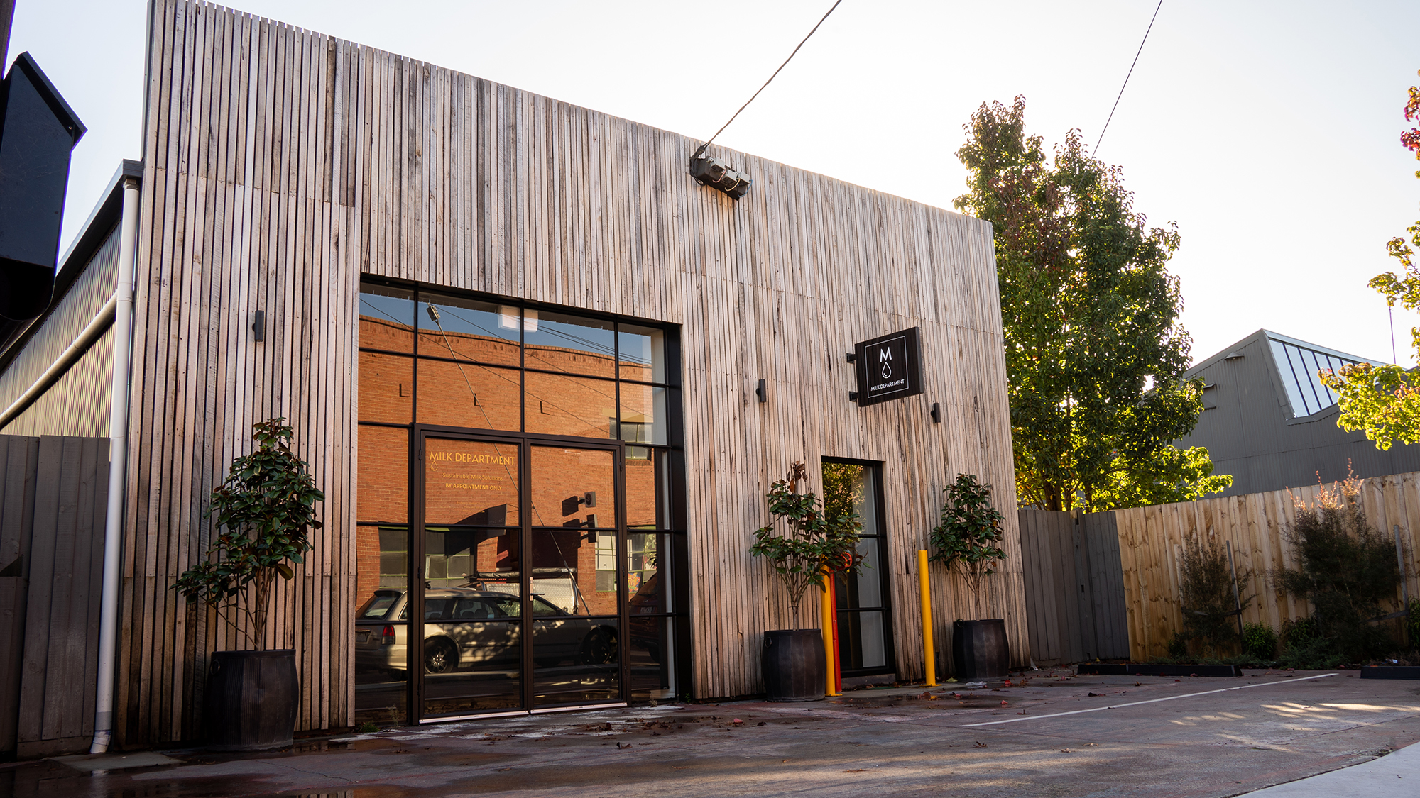 Long timber sticks run up the face of this building, in the centre is some tbar windows and a steel door, in between the doors are some small trees in black pots and in front of the building is a small carpark, Building completed by Hamilton Group Construction