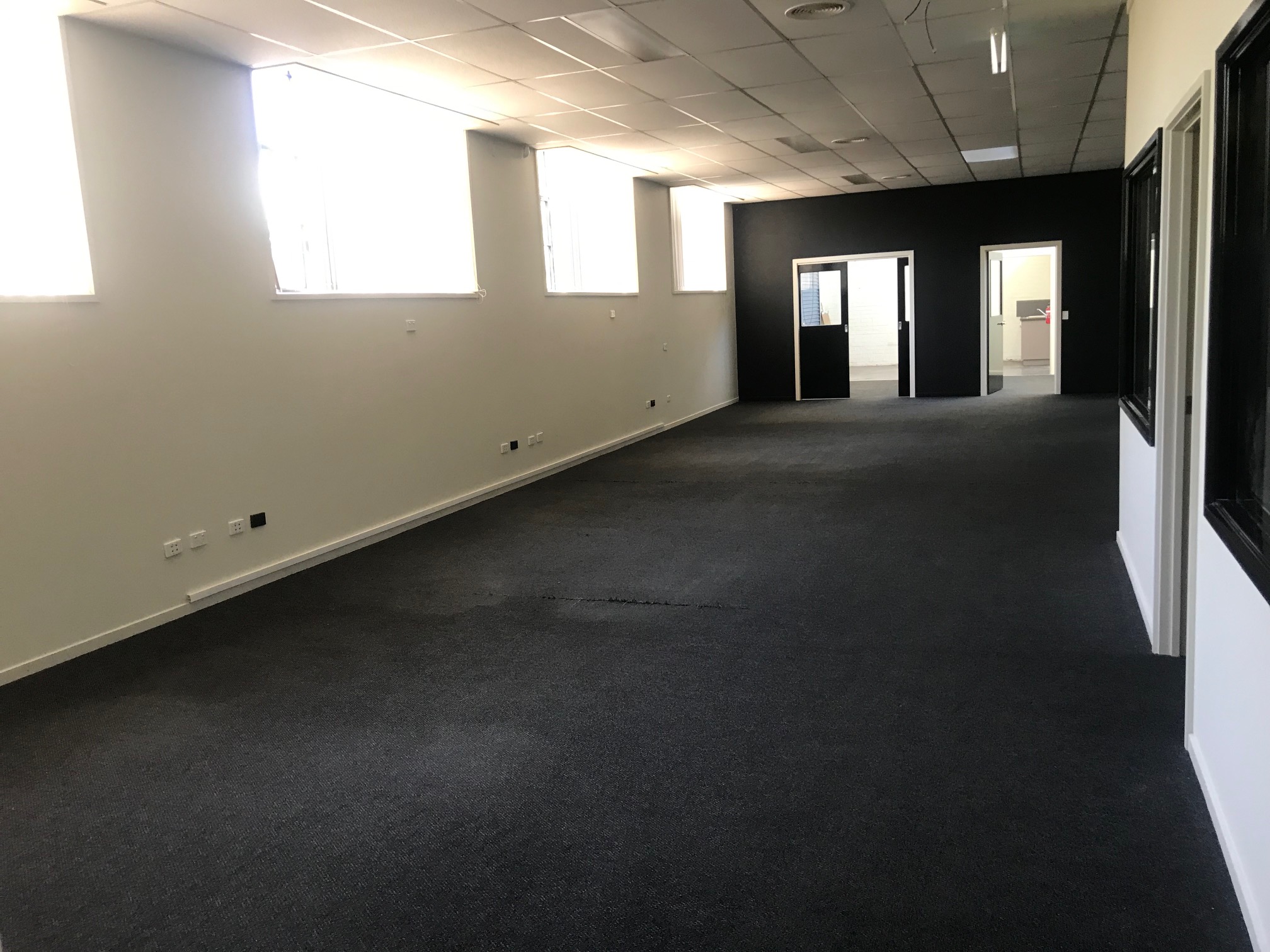 A dark office space with dim lighting and a dark carpet, creating a cozy, subdued atmosphere. The left side of the room features large windows, letting in some natural light, which contrasts with the otherwise moody ambiance of the space.