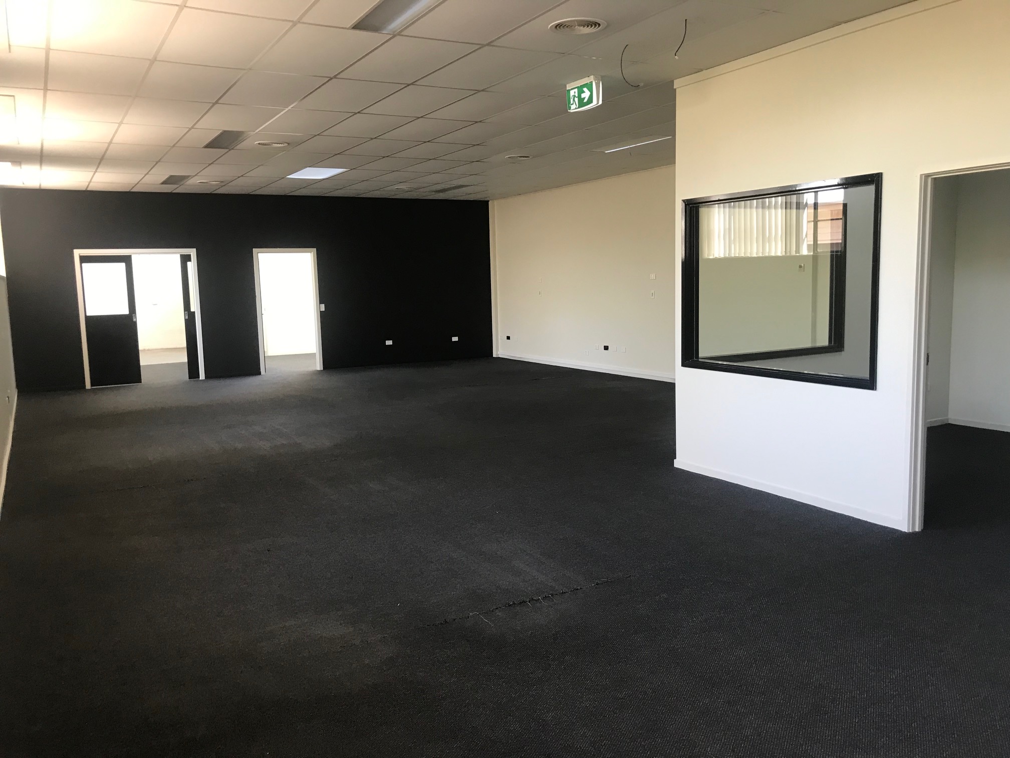 A modern office space with dark carpets and black double doors at the end of the room. To the right, there is a small office with a glass partition, adding a sense of openness while maintaining privacy. The space has a professional and contemporary feel. Hamilton Spaces