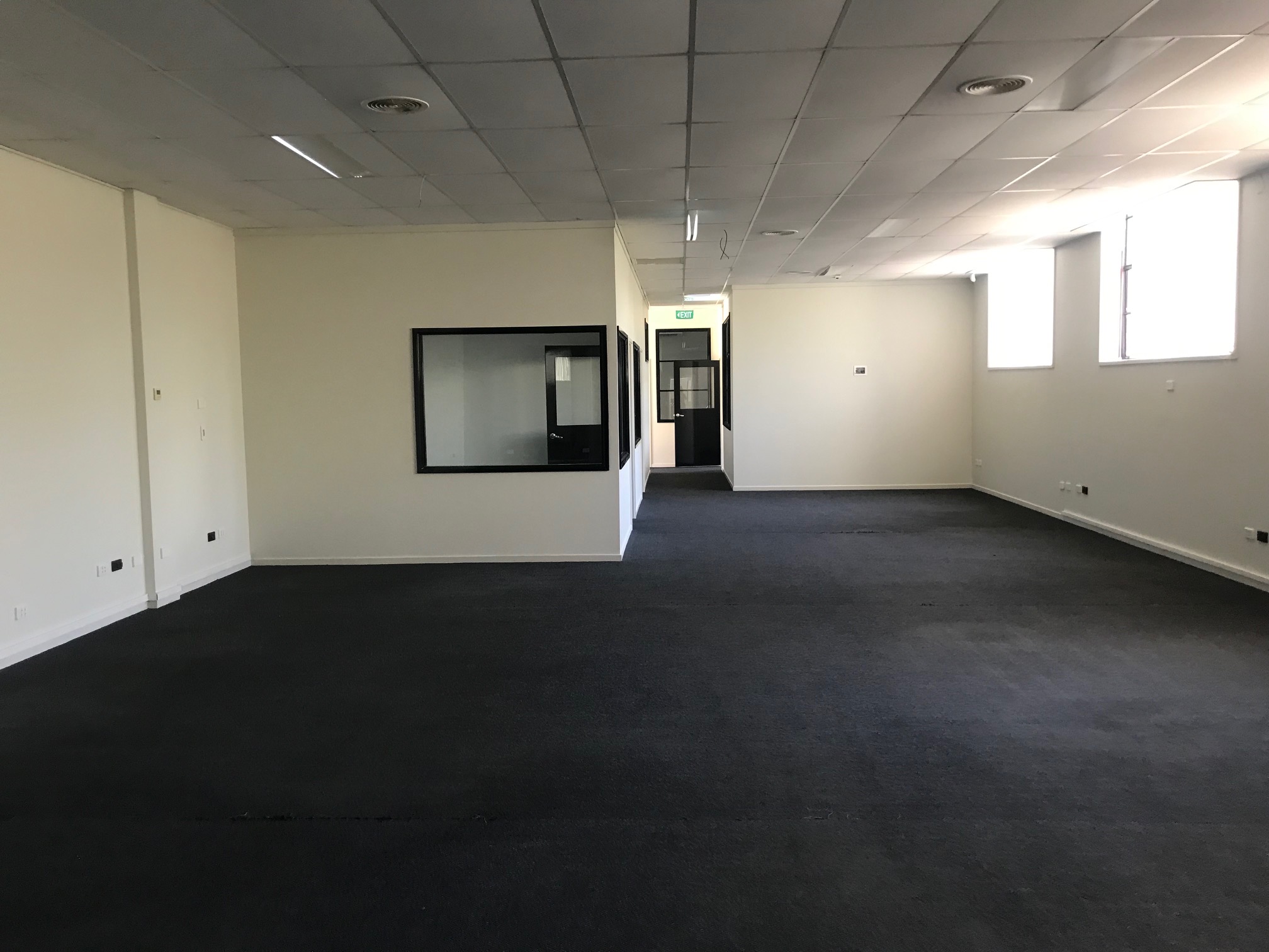 Small dark office space with dark blue carpet running throughout, in the back left corner in an even smaller office with windows opening out, two small windows to the right let as much natural light in as possible.