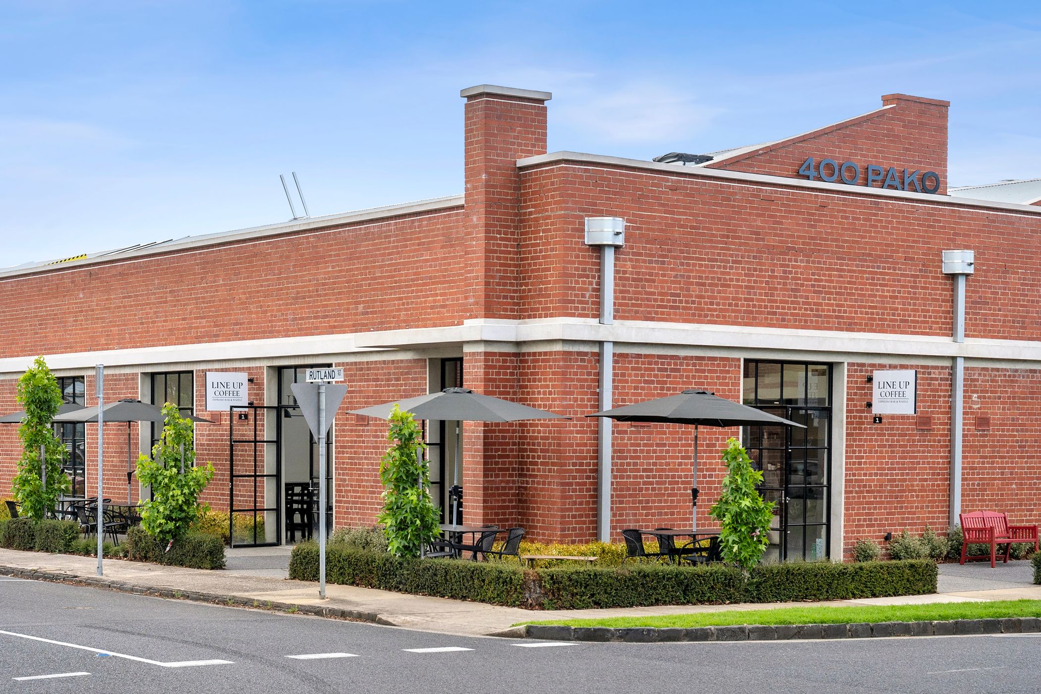 Corner view of a large redbrick facade building with a sawtooth roof, steel letters along the roof line spell "400 PAKO" black T-bar windows are spread through the facade, four large black umbrellas are stood through small black tables out the front of the building, Woolstores Geelong