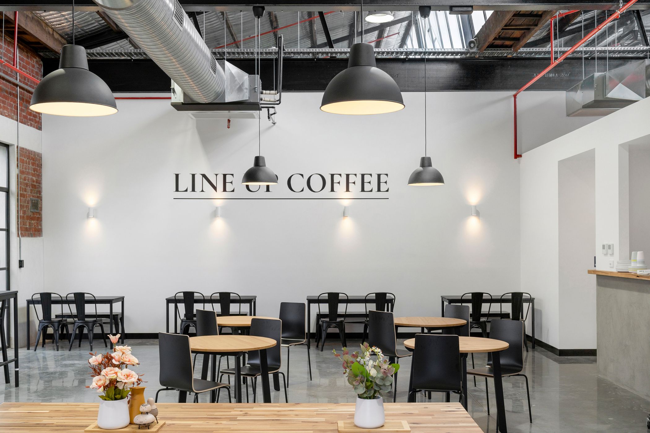 Modern cafe space using light and dark colours to create a warm and inviting space, round timber tables with black chairs surrounding them, red brick wall to the left and a concrete counter to the right, Line Up cafe within the woolstores precinct, Hamilton Group Geelong