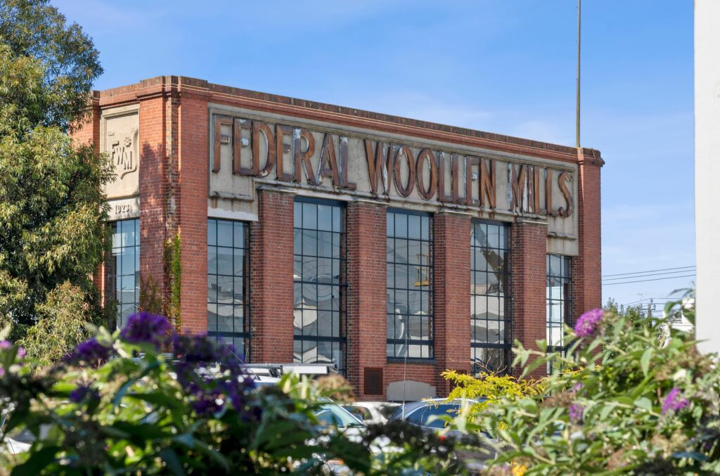 Alt text: A historic red-brick building with large industrial-style windows and an aged sign reading "FEDERAL WOOLLEN MILLS" in raised lettering on its facade. The structure features decorative concrete details and an emblem with the initials "FWM" on the side. In the foreground, green foliage and purple flowers partially frame the building, while parked cars and power lines are visible in the background. The sky is clear and blue
