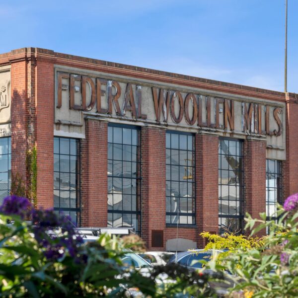 Alt text: A historic red-brick building with large industrial-style windows and an aged sign reading "FEDERAL WOOLLEN MILLS" in raised lettering on its facade. The structure features decorative concrete details and an emblem with the initials "FWM" on the side. In the foreground, green foliage and purple flowers partially frame the building, while parked cars and power lines are visible in the background. The sky is clear and blue
