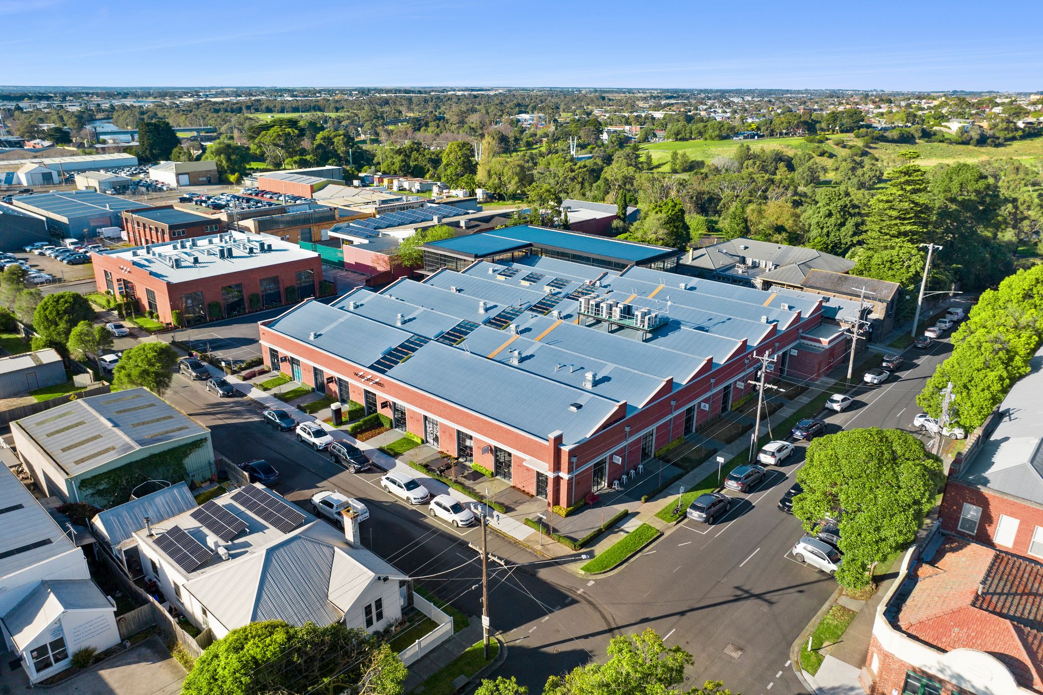 A large red-brick industrial-style building with a modern metal roof, featuring skylights and HVAC units. The building is located on a corner lot with wide streets lined with parked cars and green trees. Surrounding the structure are smaller residential and commercial buildings, some with solar panels. In the background, there is a mix of industrial facilities, office buildings, and lush green open spaces. The sky is clear and blue, with sunlight casting shadows on the streets and rooftops, Woolstores Geelong
