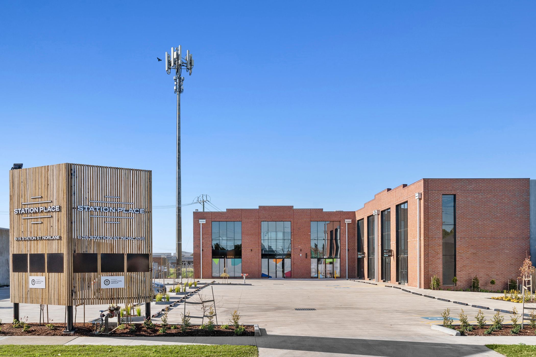 Street view of a red brick building, tall tbar windows line the front of the buildings, large concrete car park featuring multiple trees in the front and a large steel and timber sign on the left with "Station Place" on it, Hamilton Property Group Geelong