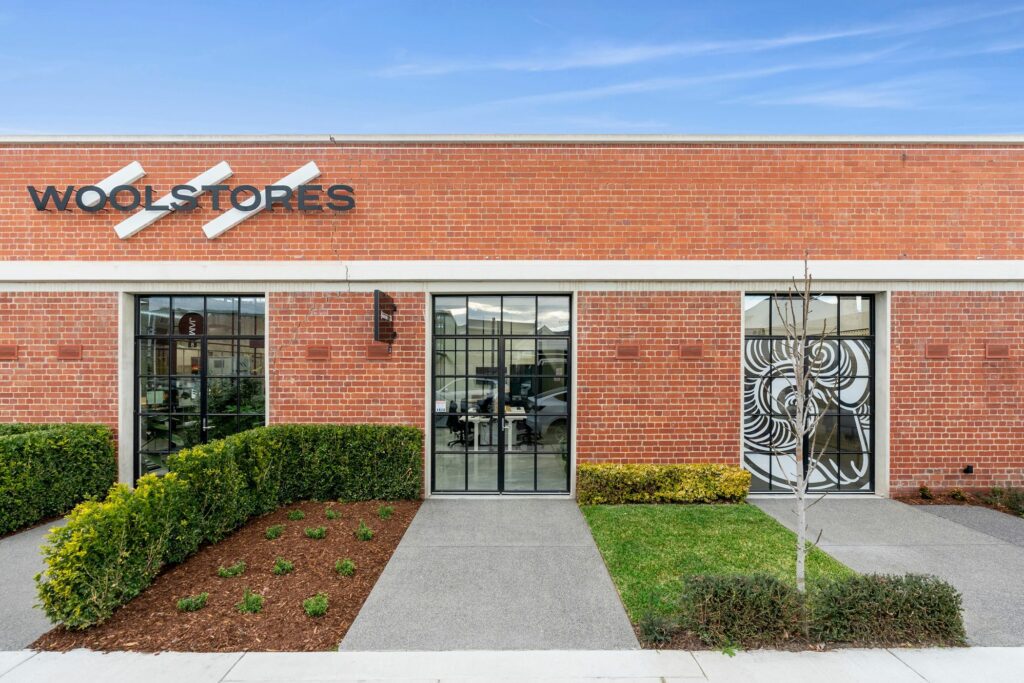 Red brick facade stretches across the shot, well maintained lawns fit between concrete pathways, black T-bar windows and doors and spread across the building, large "woolstores" sign attached to facade, decal of a ram on the window, Gardens completed by Hamilton Landscaping