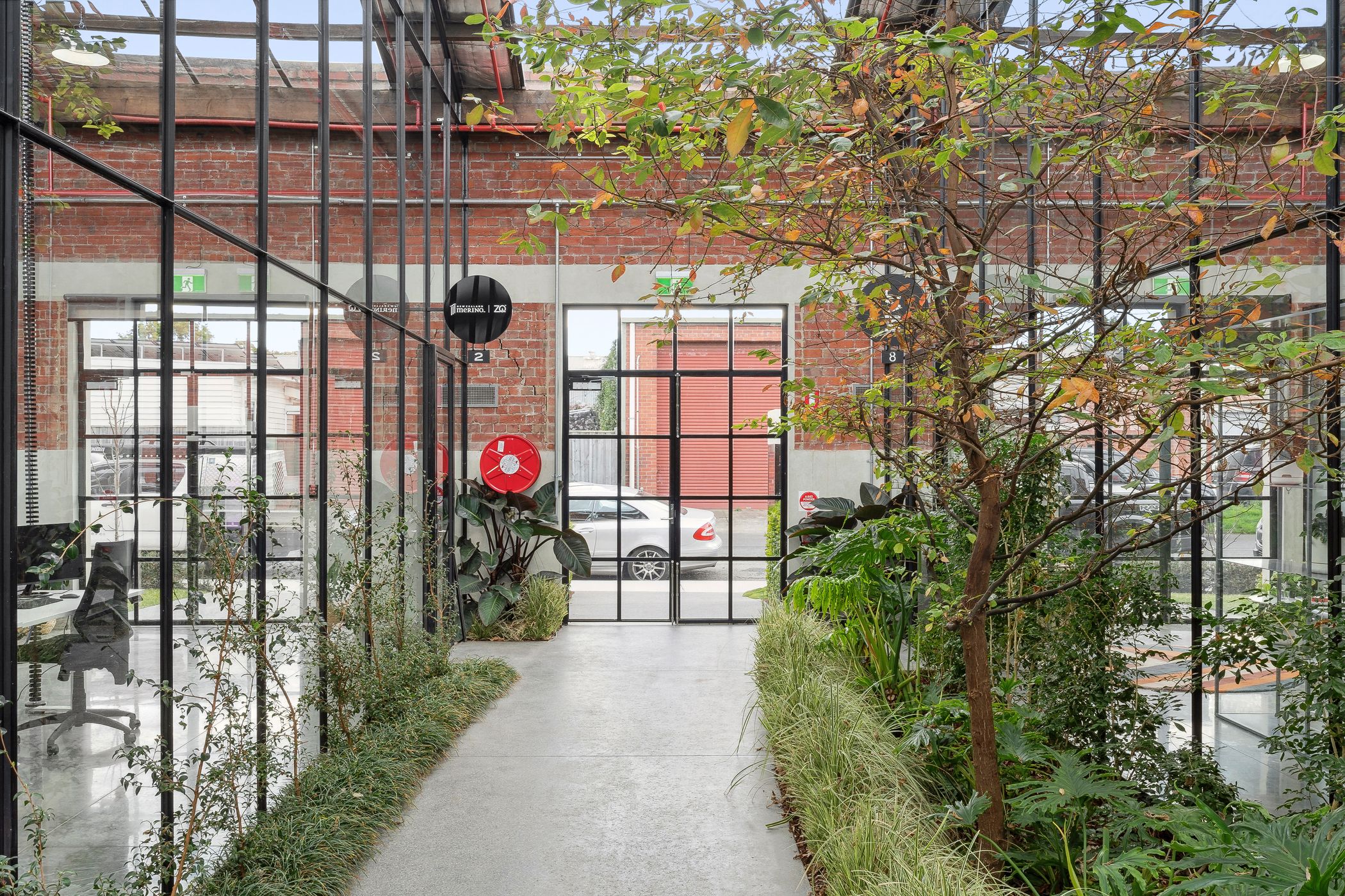 Pathway running down the centre, leading to t-bar double doors and windows, lush green plants and trees line the edges of the path, a red brick wall sounds the windows and doors. Hamilton Property Group Woolstores