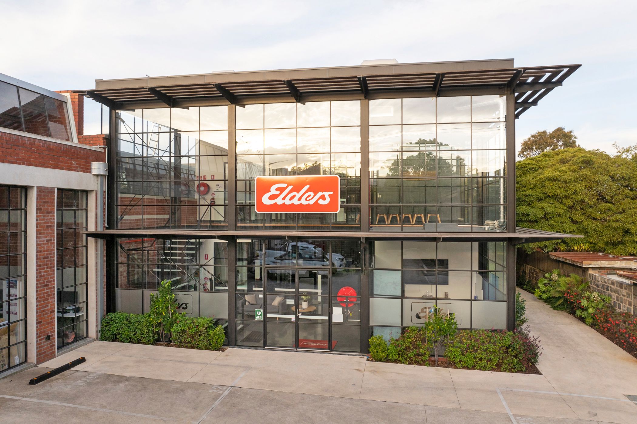 Large black building filled with floor to ceiling T-bar windows and doors, a heavy timber awning surrounds the structure, lush green plants are around the base, a concrete carpark is out the front and a footpath is leading off behind, located at woolstores Geelong