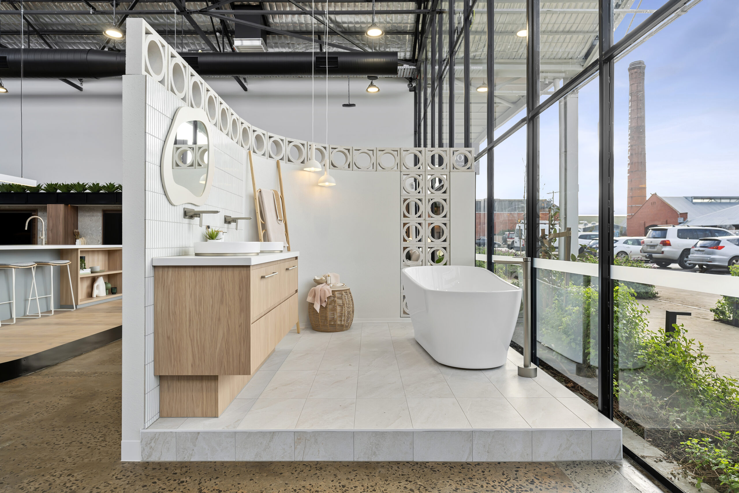 Internal view of bathroom showroom, elevated platform with tiled floor, vanity with taps and a sink, a bathtub and a petition wall to separate each area. View through T-bar windows to carpark and large red brick chimney in the distance, Hamilton Group Approach.