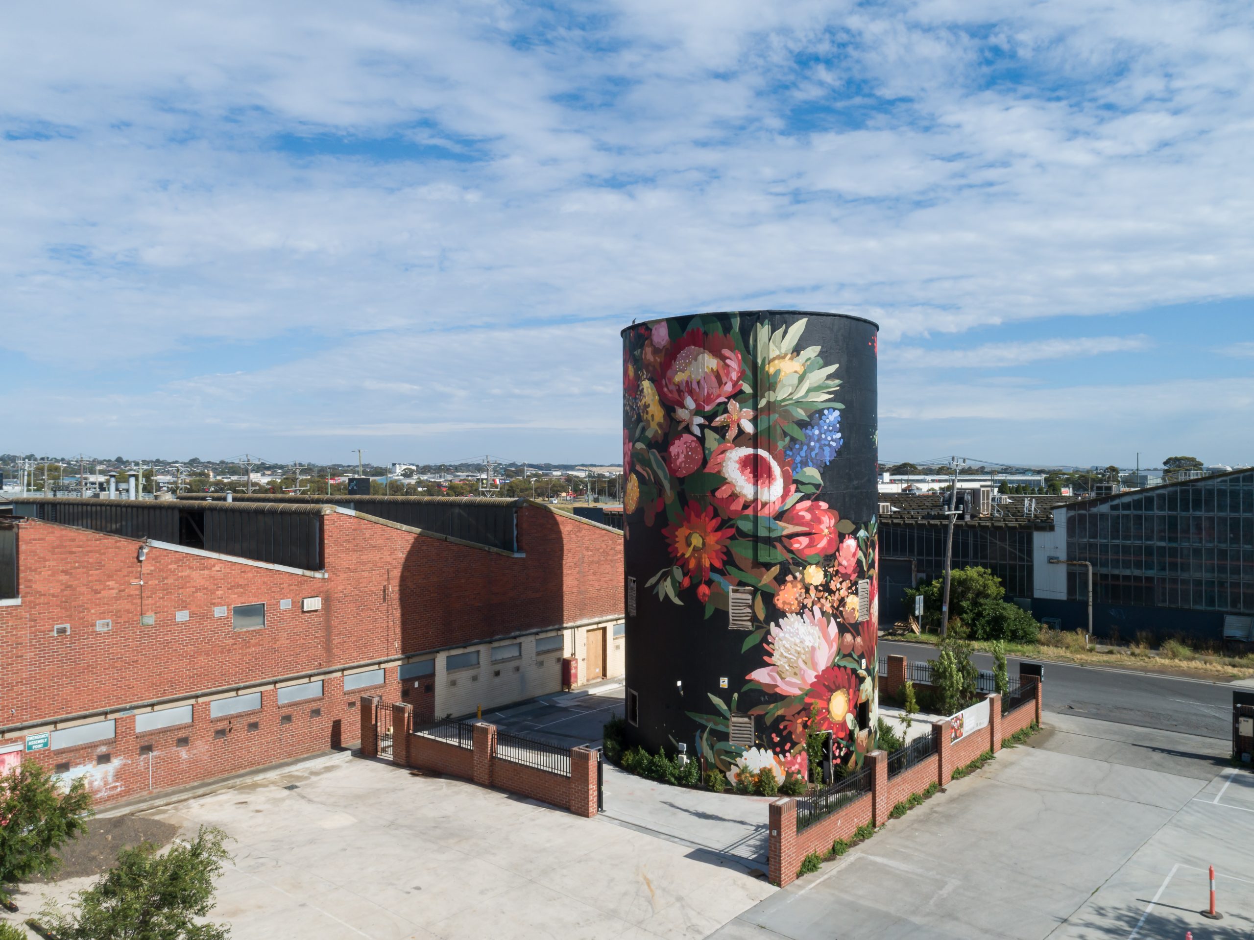 A large cylindrical silo at Federal Mills in North Geelong features a striking mural by artist Sai Neoh. The artwork depicts a vibrant floral arrangement with red, pink, yellow, and blue flowers set against a black background. The silo is surrounded by industrial brick buildings, with a clear blue sky and distant hills in the background.