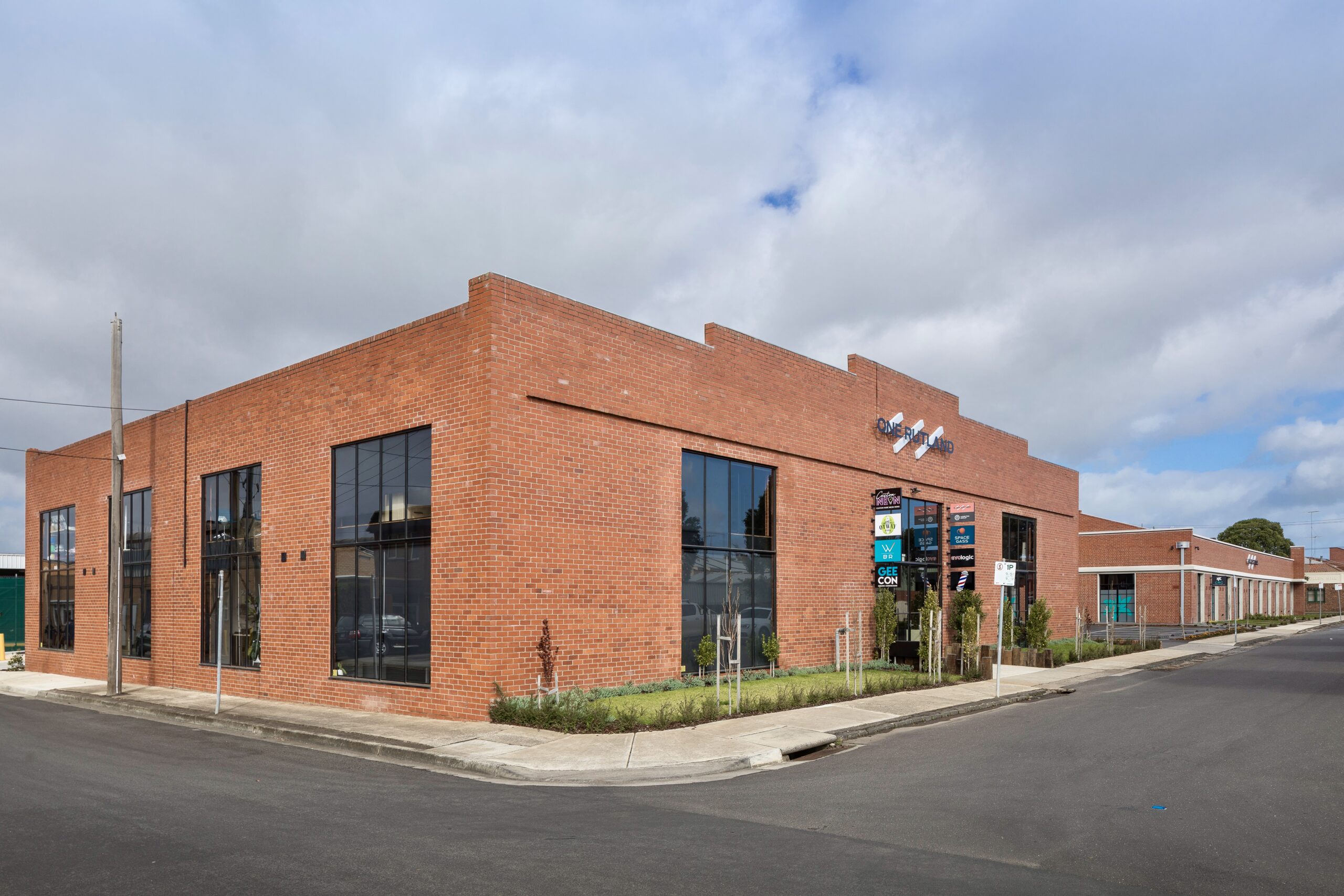 Large red brick establishment with tall windows scattered around, large sign on the front saying "ONE RUTLAND" below that in eight signs with different logos, a small garden lines the front of the building, Hamilton Group Geelong