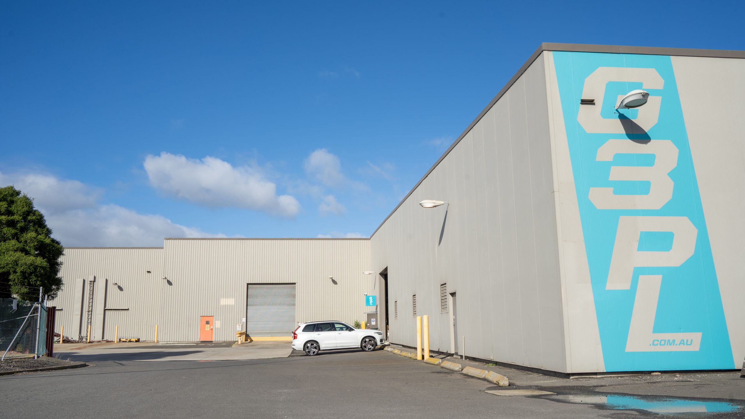 Carpark in the front with one white car parked, large white shed surrounds the carpark with multiple garage doors, large blue sign with "G3PL" painted on the front, Owned by Hamilton Property Group Geelong