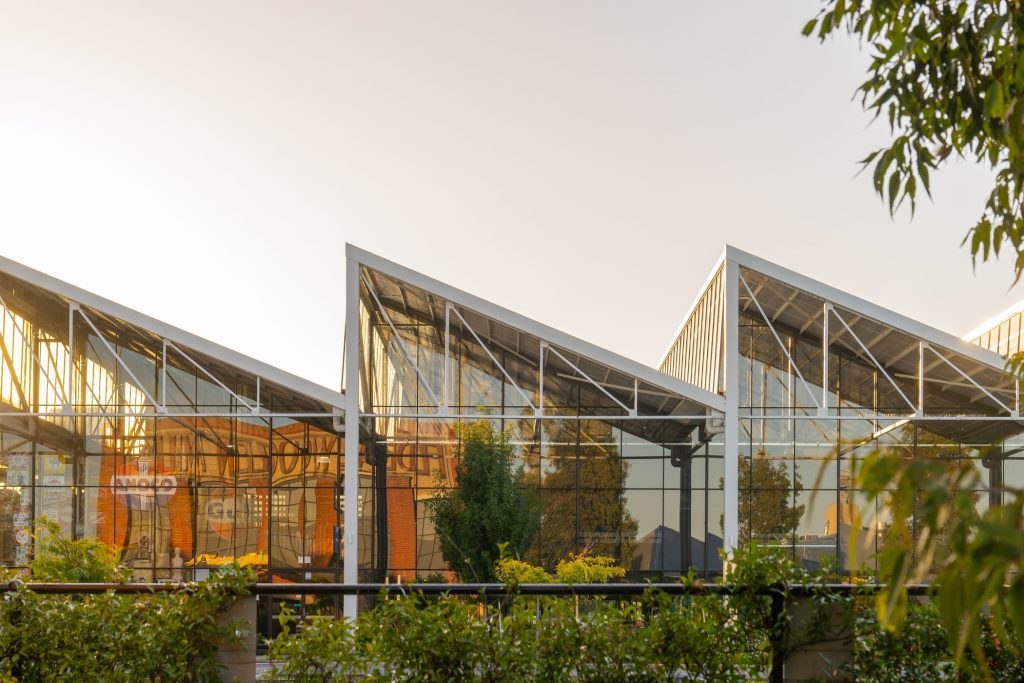 "A modern glass building with angular, slanted rooftops supported by white steel frames. The reflective glass panels showcase the surrounding greenery and sky, with warm sunlight casting a golden hue on the structure. The interior, partially visible through the transparent facade, includes signage and decor. Lush green plants frame the foreground, adding to the natural ambiance.
