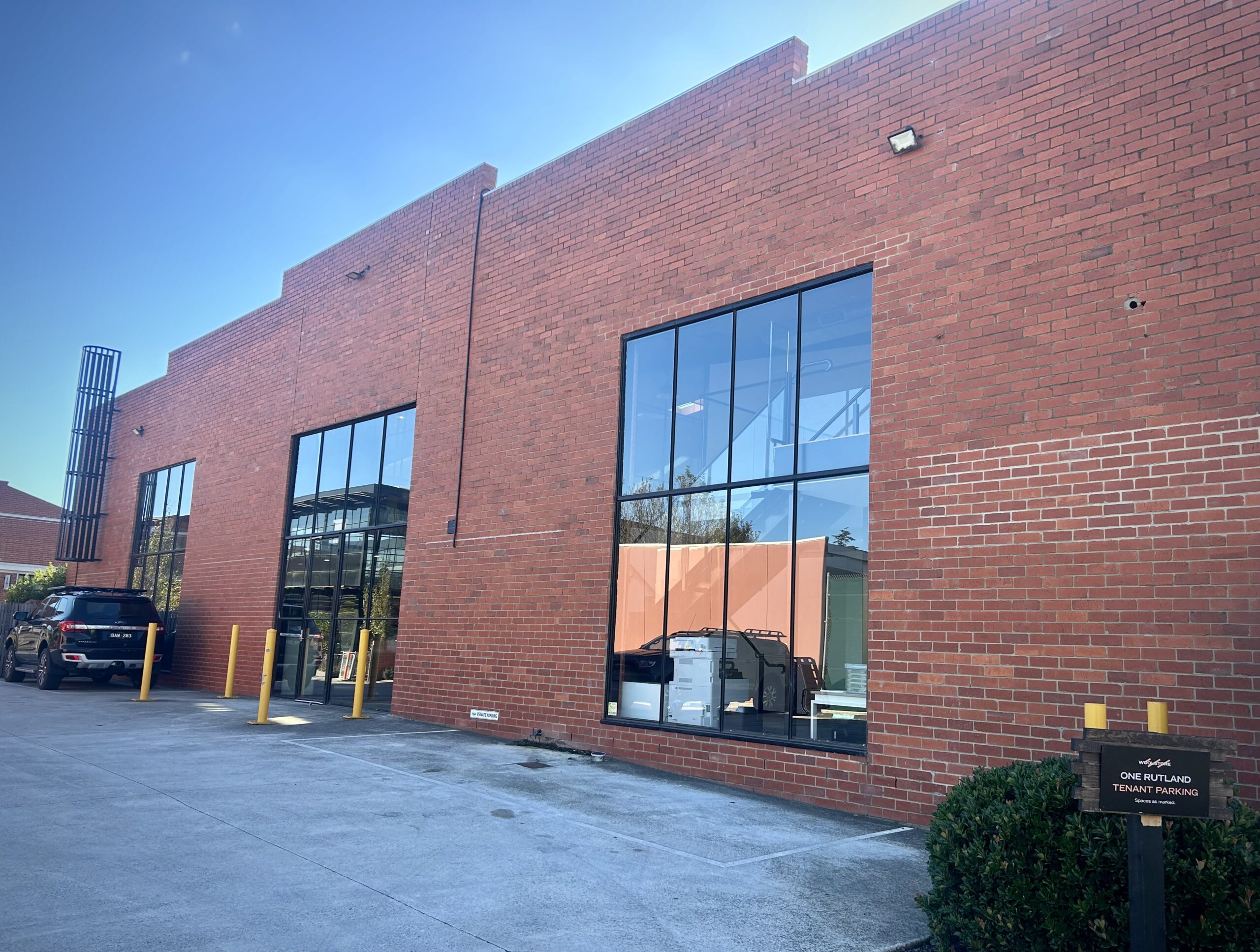 Tall red brick facade with tbar windows spread throughout, a door way in the centre is protected by four yellow bollards, a concrete carpark is in the front of the shot featuring one black car, Hamilton Group Geelong