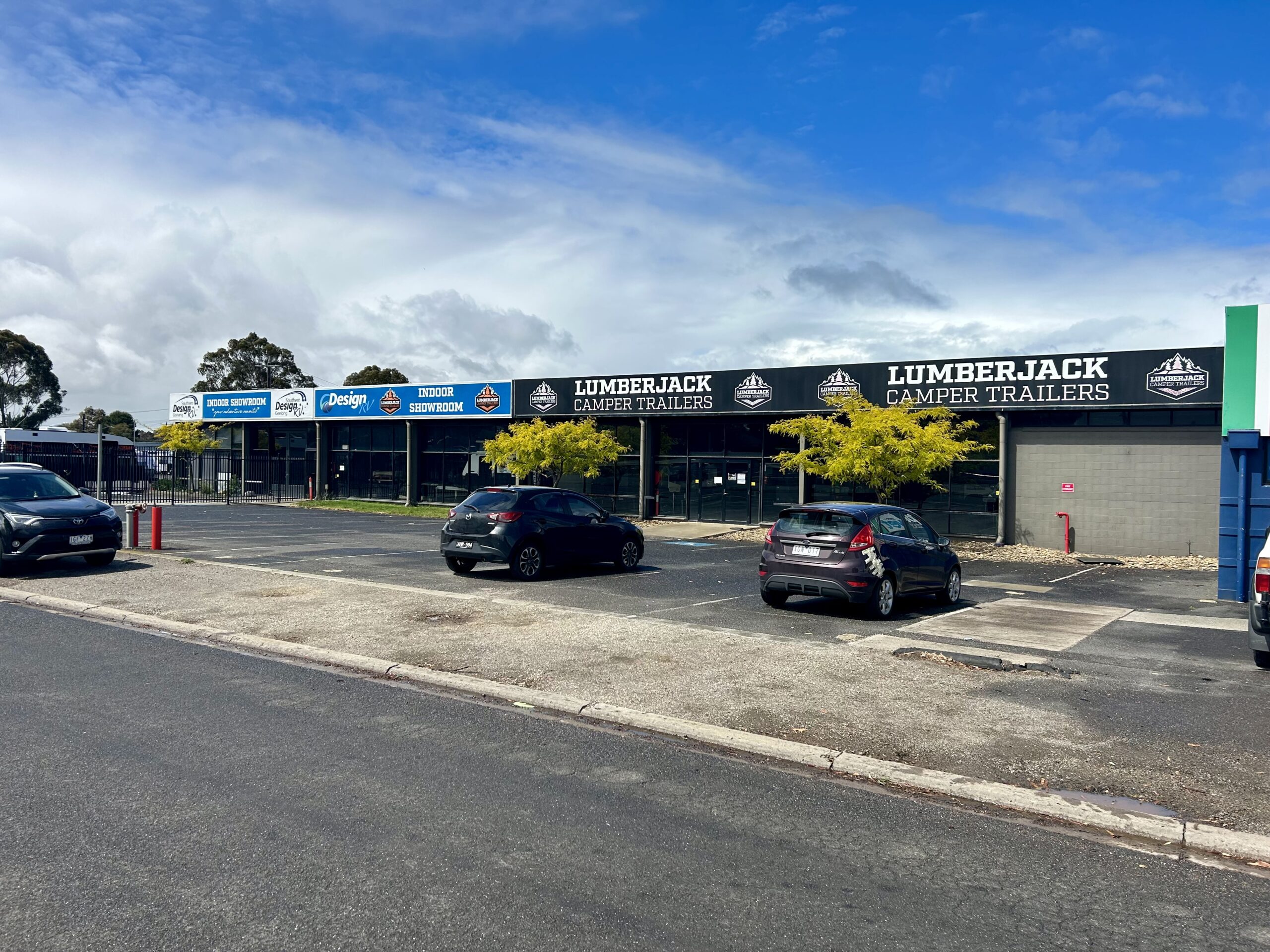 Cloudy blue sky above, old concrete and asphalt carpark in front of building featuring three parked cars and some yellow trees, buildings have long windows running across the span and theres some water hydrants scattered around, Building owned by Hamilton Group Geelong