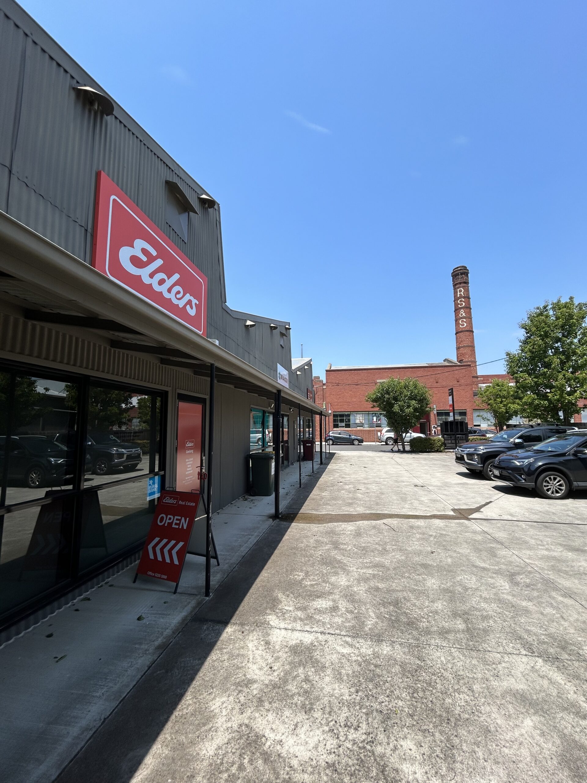Clear blue skies overhead, tall red brick chimney in the distance, old grey concrete carpark in the centre of shot, two cars are parked to the right side, lush green trees surround the carpark, Hamilton Geelong properties