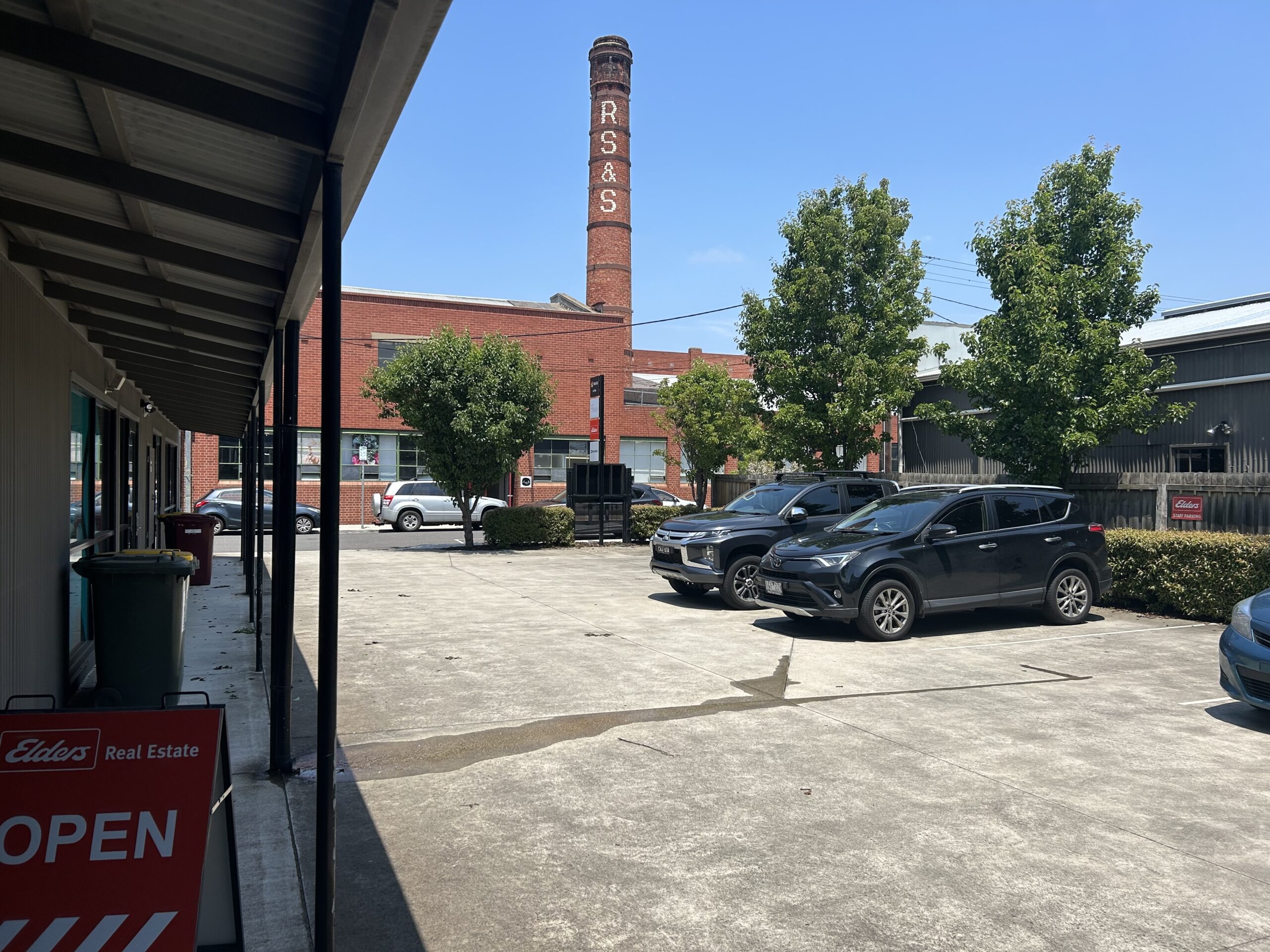 Clear blue skies overhead, tall red brick chimney in the distance, old grey concrete carpark in the centre of shot, two cars are parked to the right side, lush green trees surround the carpark, Hamilton Geelong properties