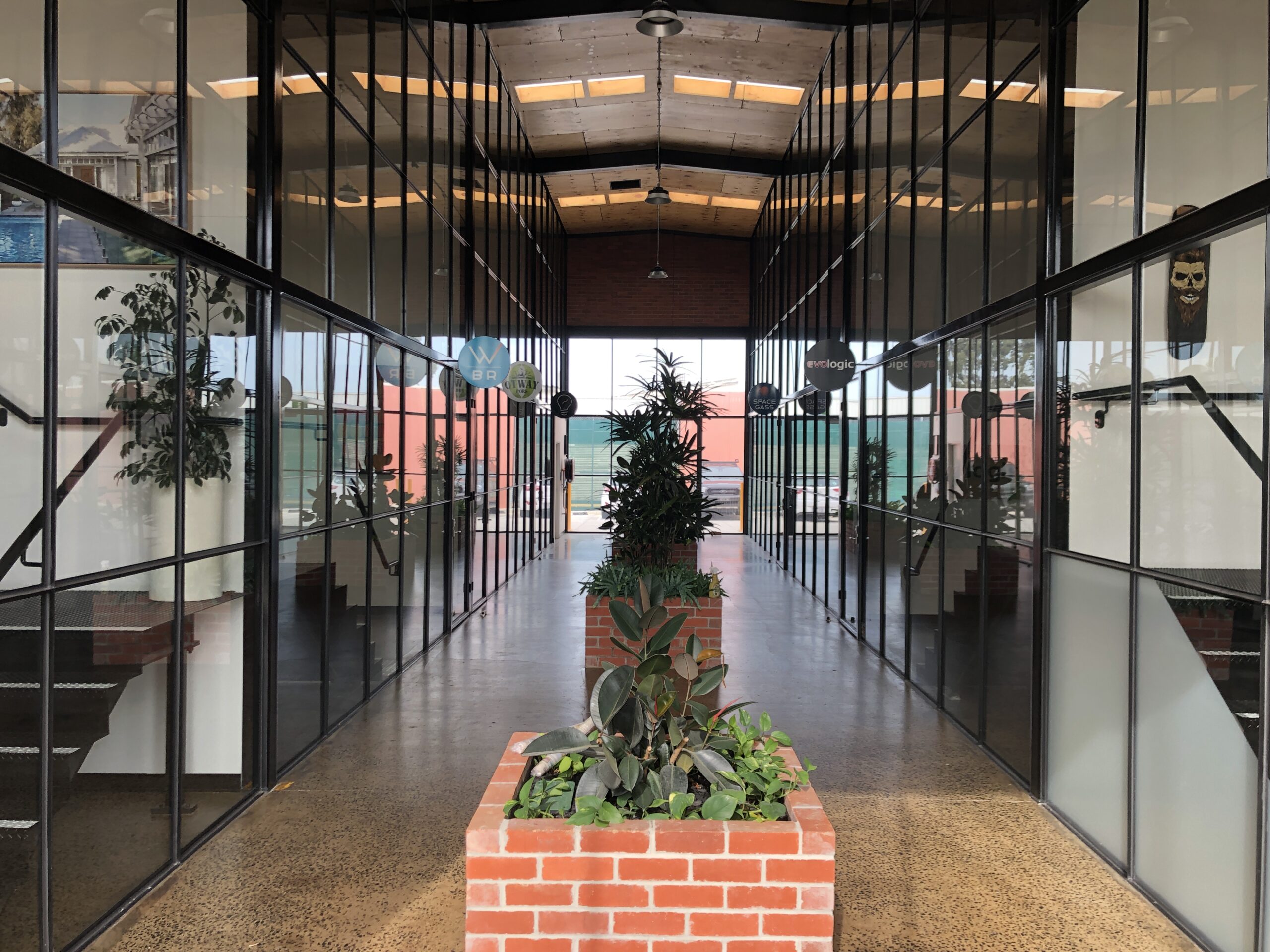 Large industrial shed converted into individual tenancies using floor to ceiling tbar windows and doors, polished concrete floors, three red brick planter boxes filled with plants, Hamilton Group Geelong