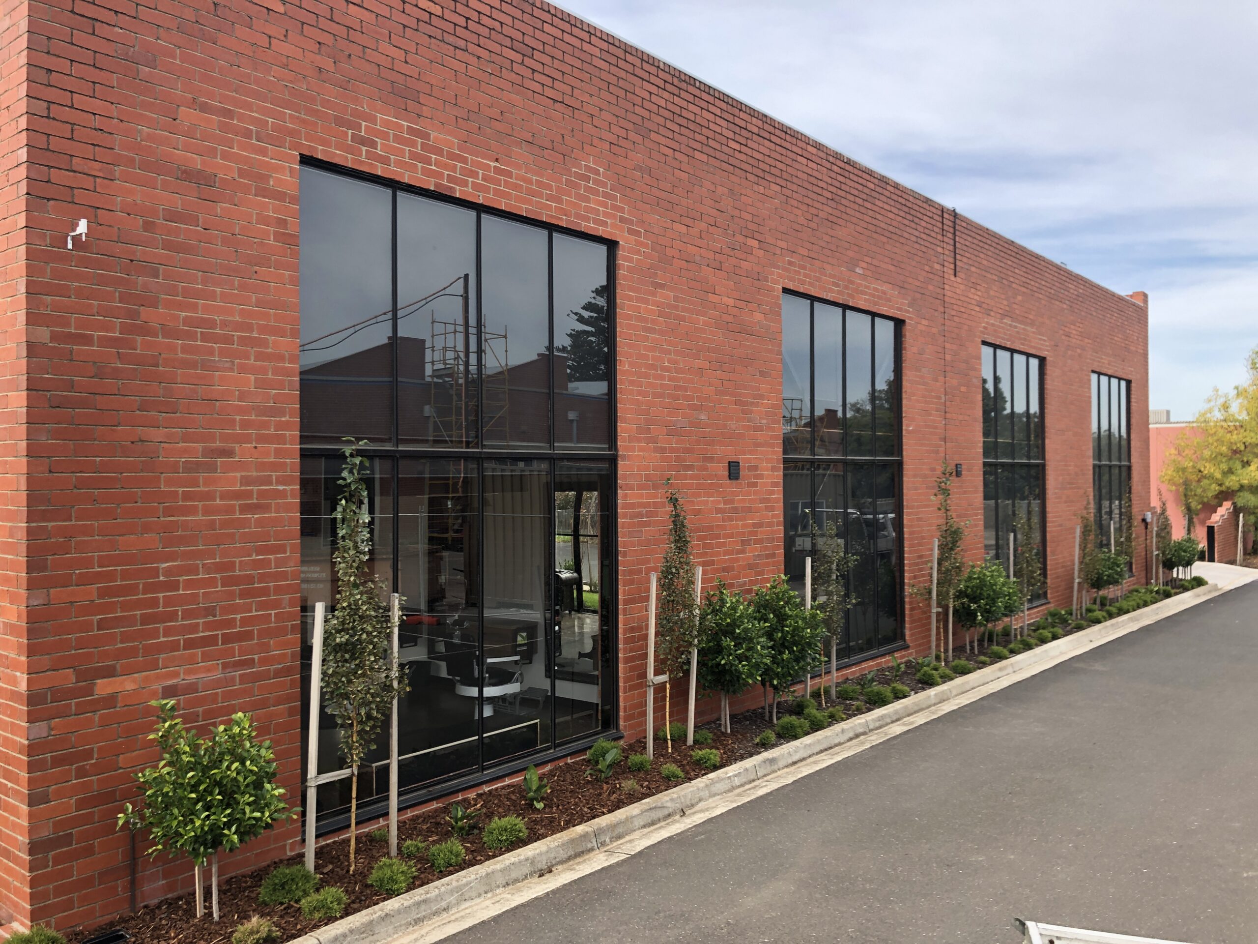 Red brick facade with four large tbar windows , cloudy sky above and a lush green garden in front and a concrete driveway leading past the building, Hamilton Property Specialists