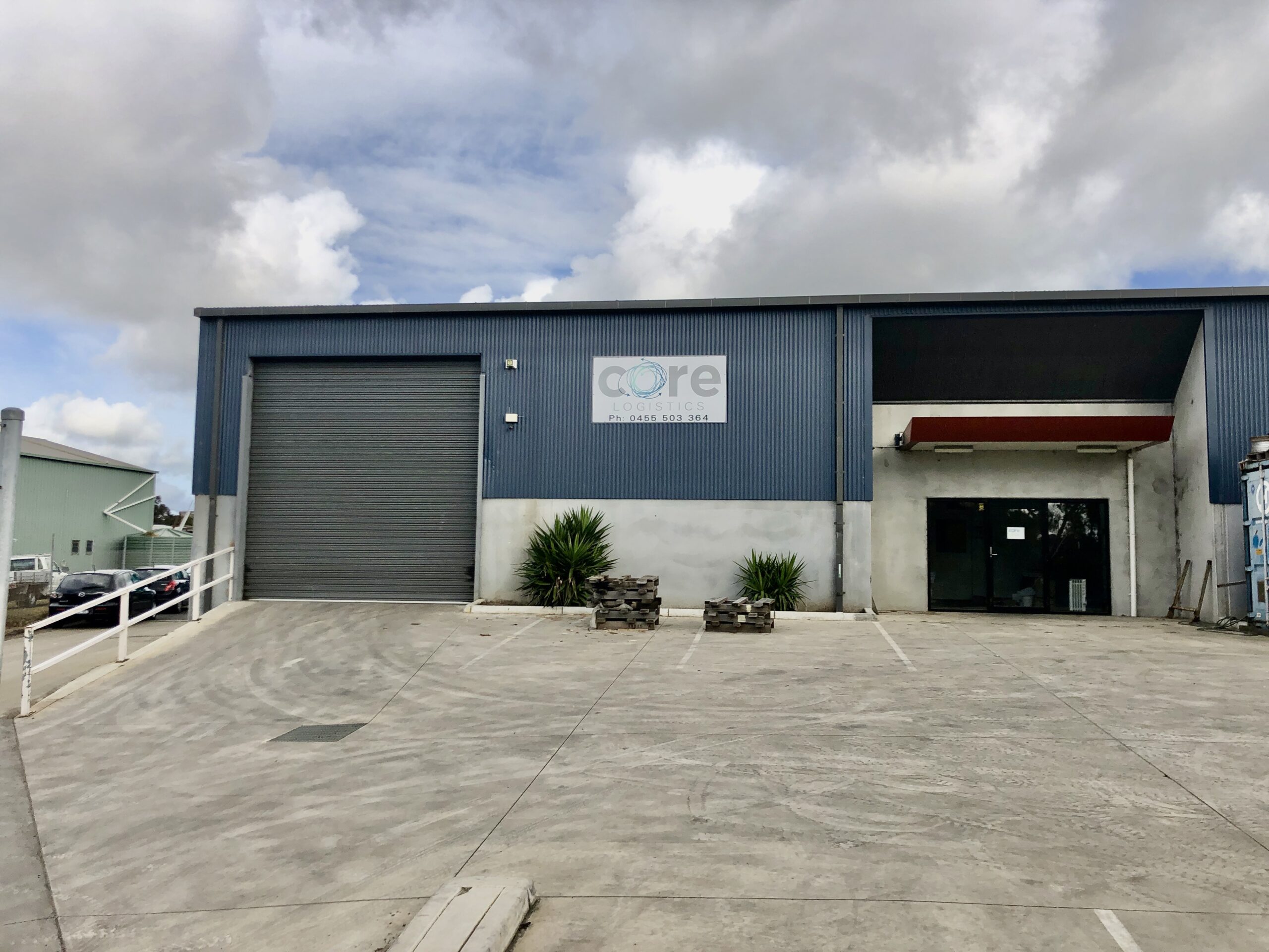 Concrete car park covered in tire marks leads up to large shed, the shed has a tall roller door on the left side and a front pedestrian door on the right, above the door is a small awning, cloudy skies above, Hamilton Group Geelong