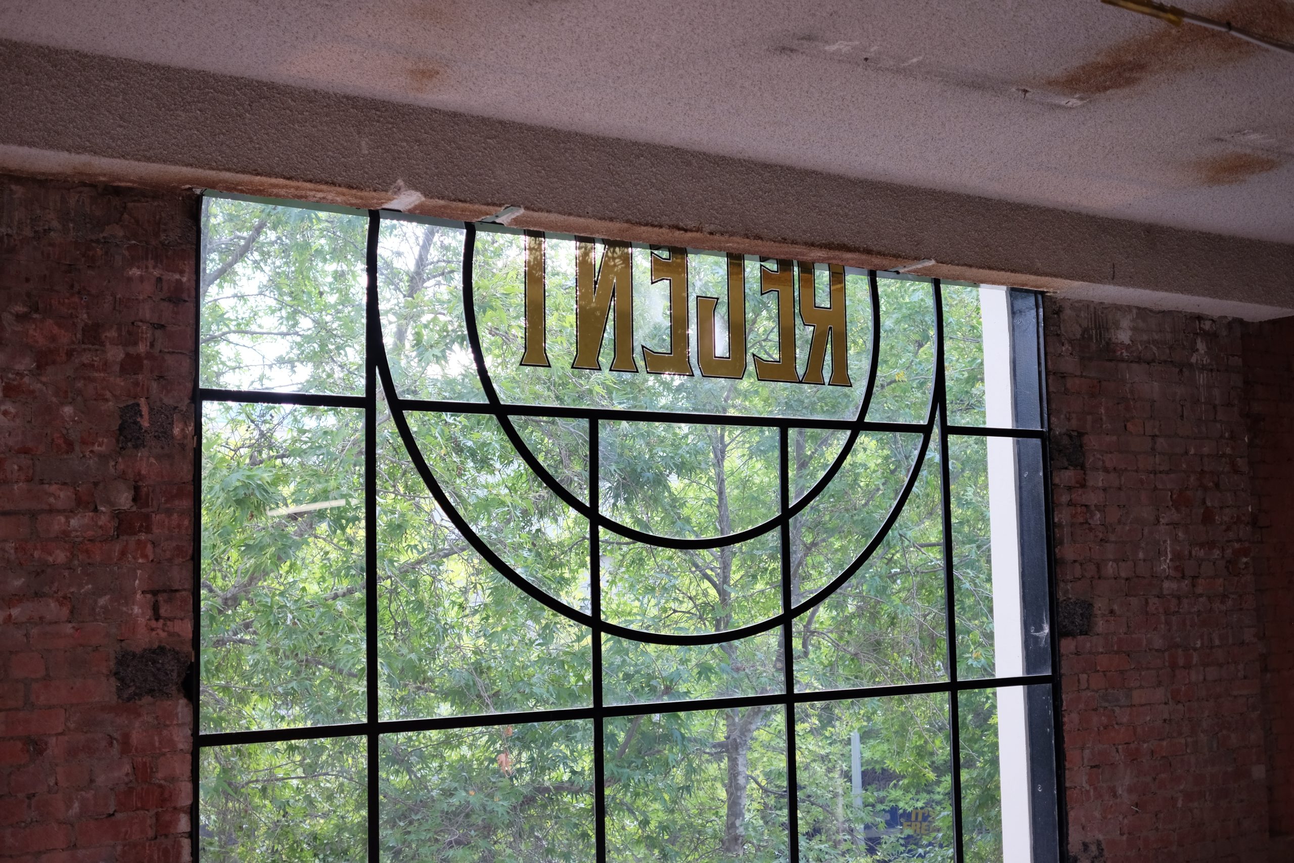 Large glass windows with fabricated tbar throughout, two round sections surround the word "REGENT" through the glass window is many lush green trees, either side of the windows are red brick walls, Hamilton Group Geelong