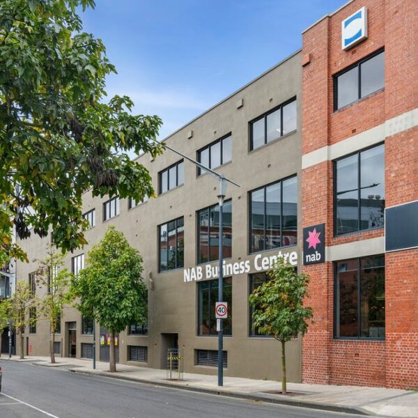 Large triple level building, dark in colour and has a corner feature of red bricks, the building is filled with many windows, a street runs across the front of the building lined with trees. Hamilton Property Group