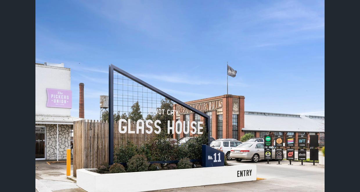 Entry way to a car park, on the left is a large fabricated steel sign with "GLASS HOUSE PIVOT CITY" welded to the front, behind the sign is the Pickers union cafe and behind to the right is the famous Federal Woolen Mills building and on top of the building is a large black flag, clear blue skies over-head, Hamilton group Geelong