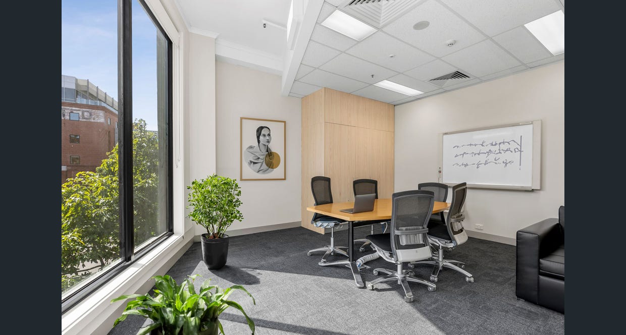 A modern, well-lit meeting room with a large window offering a view of trees and a brick building. The space features a round wooden table surrounded by ergonomic office chairs, a whiteboard with abstract writing, and framed artwork of a stylized portrait. The room is decorated with potted plants and has a neutral colour palette with wooden and black accents, creating a professional yet inviting atmosphere.