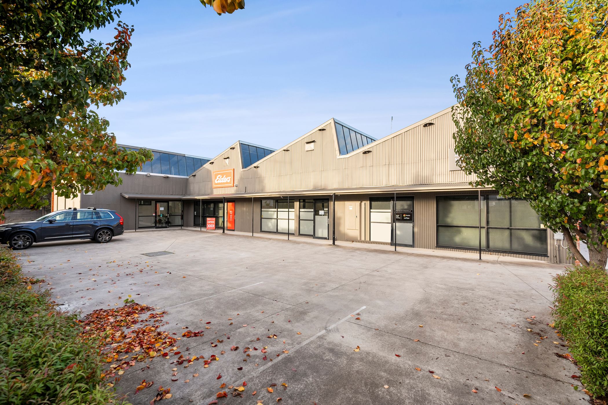 Old grey carpark surrounded by lush green trees and plants, grey shed at the back of the carpark has a sawtooth roof and clear skies above that, a single car is parked, Hamilton Group Geelong