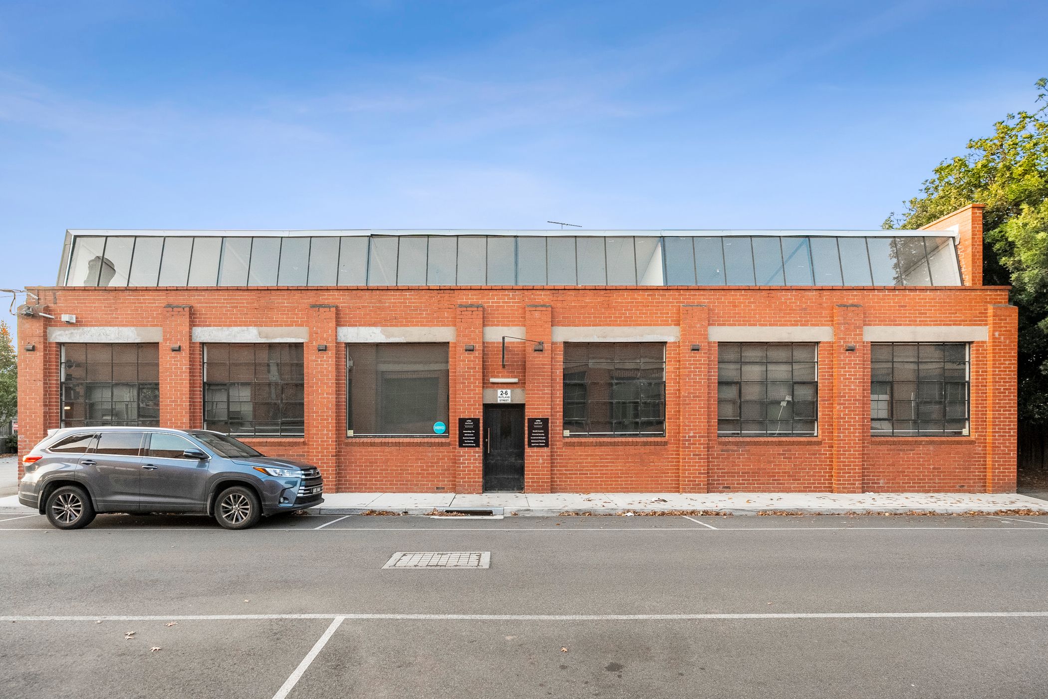 Old looking red brick facade features multiple black windows and a small black door in the centre, clear blue skies above, a roadway lines the front of the building with a single car parked out the front - Hamilton Property Specialists