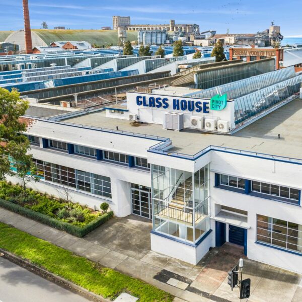 An aerial view of a white, two-story modernist building with blue accents, labeled 'GLASS HOUSE' with a 'Pivot City' sign. The building features large windows and a rooftop section with skylights. It is surrounded by trees, a sidewalk, and a landscaped garden. In the background, an industrial area with warehouses, factory buildings, and a tall chimney is visible, along with a distant view of the ocean under a partly cloudy sky.