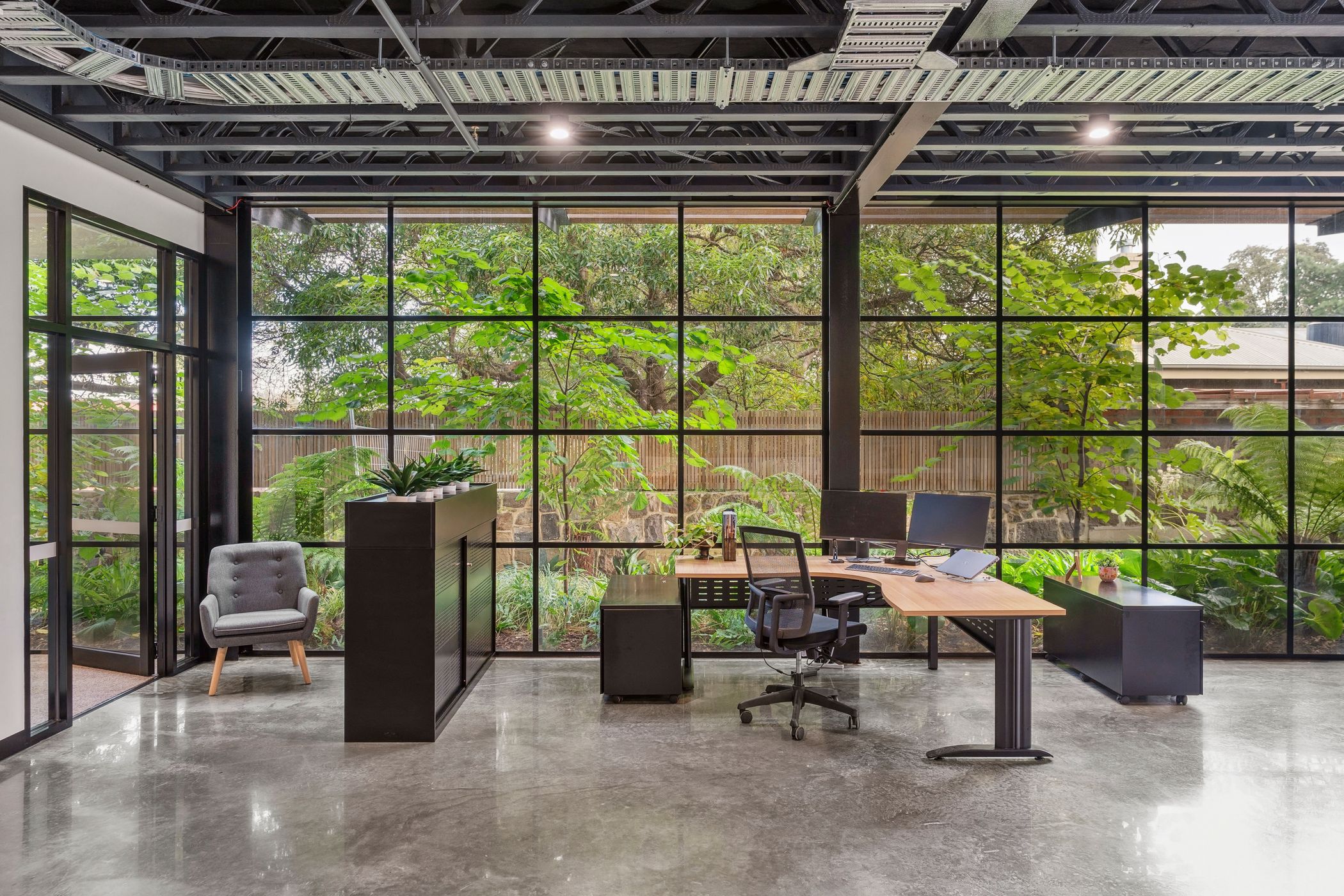 Modern office space with 'L' shaped timber desk featuring dual monitors, an ipad and an ergonomic chair, a black filing cabinet with green plants on top, a small grey chair in the corner of the room, floor to ceiling T-Bar window wall giving a view of lush green outdoor plants and trees, Woolstores owned by Hamilton Property Group Geelong