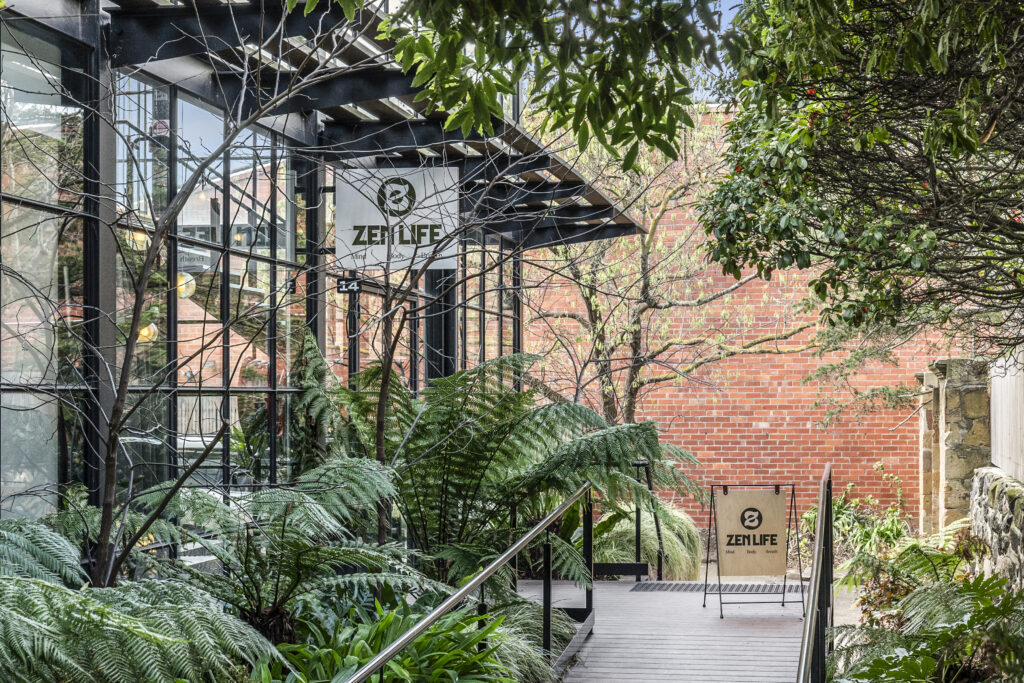 Timber ramp with black steel handrail lead up to industrial facade, fit-out with T-bar windows and doors. either side of the ramp is covered in organic green plants and trees all planted by Hamilton Landscaping.