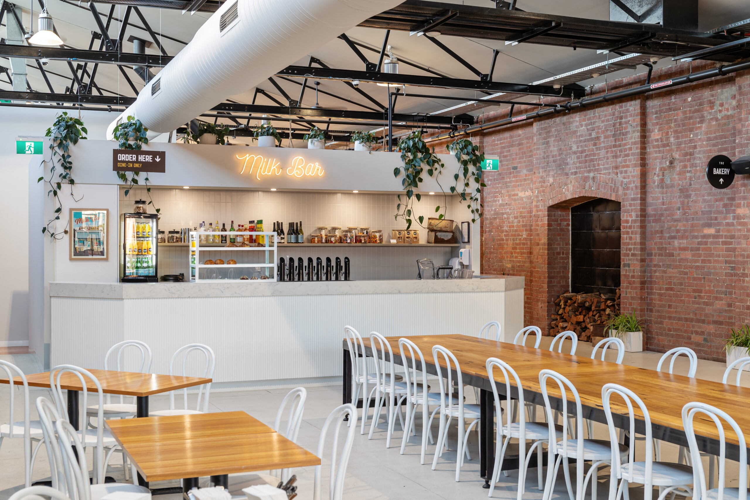 Long timber table with black steel fabricated legs, surrounded by white chairs, in the back is a bar area with multiple beer taps and a shelf full of drinks above the bar is a sign saying 'Milk Bar' Hamilton Group Geelong