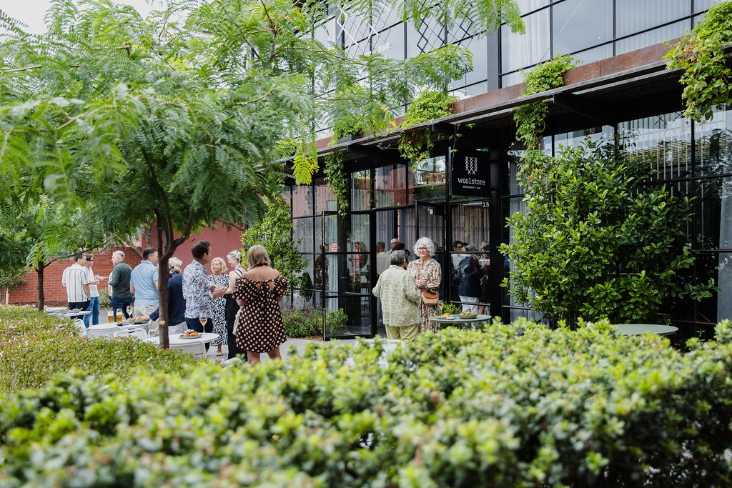 A lush outdoor gathering space in front of a modern glass-fronted building with a sign reading "Woolstores." People are socializing in small groups, dressed in casual and semi-formal attire, with drinks and food on tables. The area is surrounded by greenery, including trees, bushes, and hanging plants. The setting has a relaxed and welcoming atmosphere.