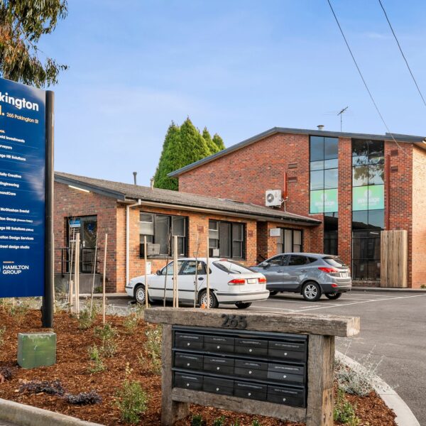 Double story red brick complex with large black windows, two cars are parked out the front, manicured gardens surround the building with a sign out the front saying "Pakington Hill" Hamilton Group Geelong