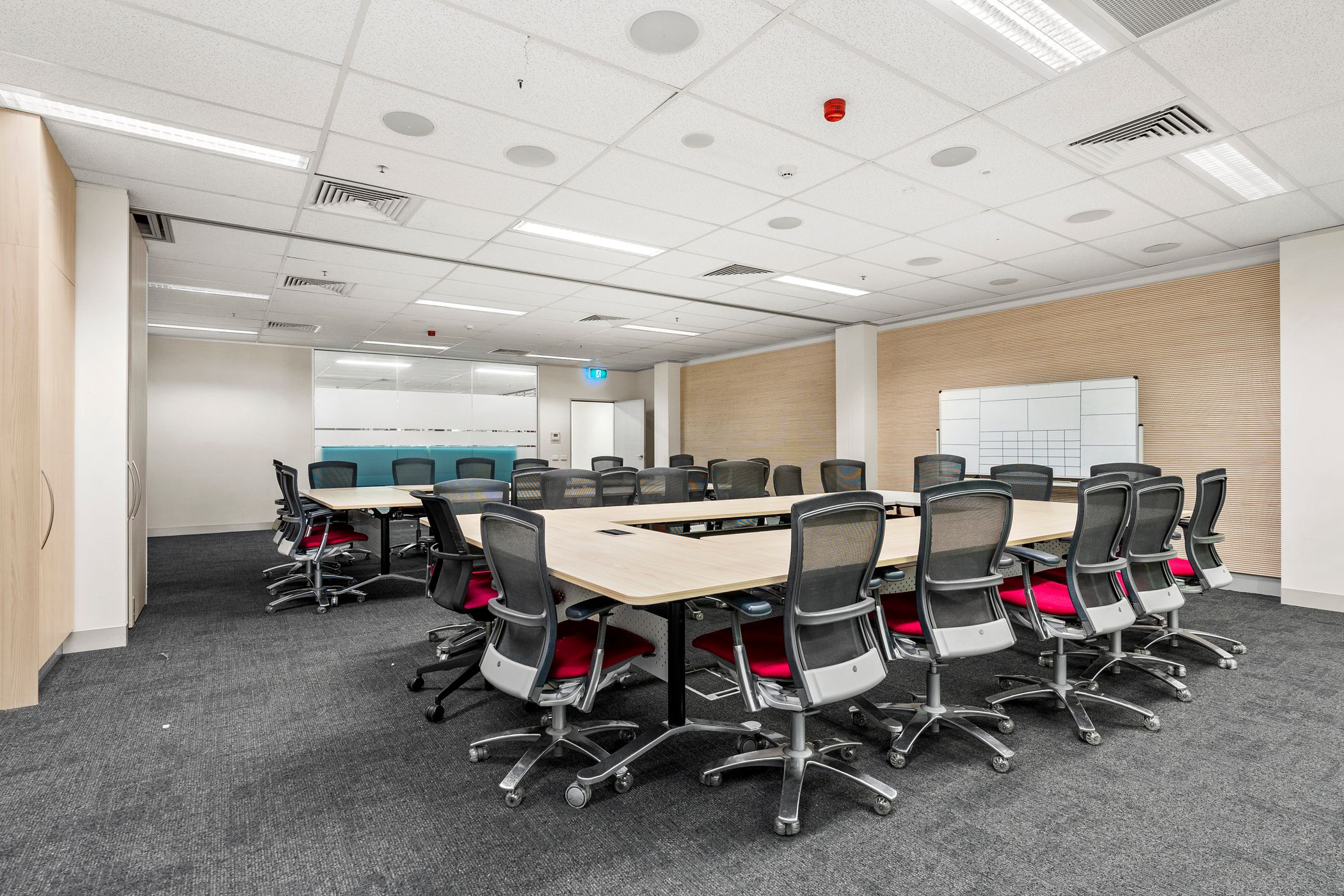 A modern conference room with a large U-shaped table surrounded by ergonomic chairs with red cushions. The space is well-lit with recessed ceiling lights and has a neutral color scheme with wooden accents. A whiteboard and storage cabinets are positioned against the wall. Glass partitions in the background provide separation from other office spaces while maintaining an open feel.