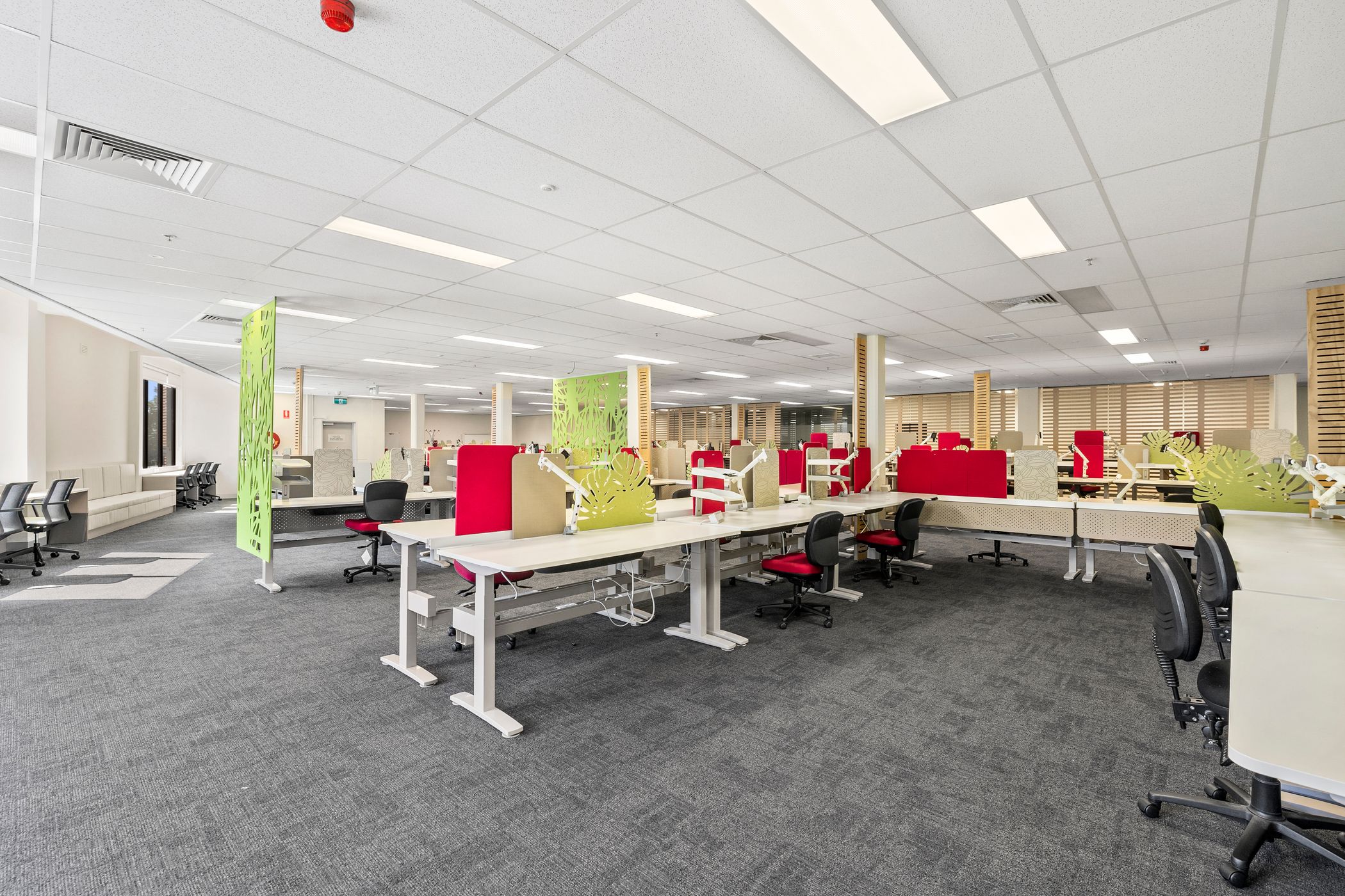 This is a modern open-plan office space with shared workstations, ergonomic chairs, and a well-lit environment. The red and black chairs contrast with the neutral decor, and there are partitions with green decorative elements. It looks like a collaborative workspace designed for productivity.