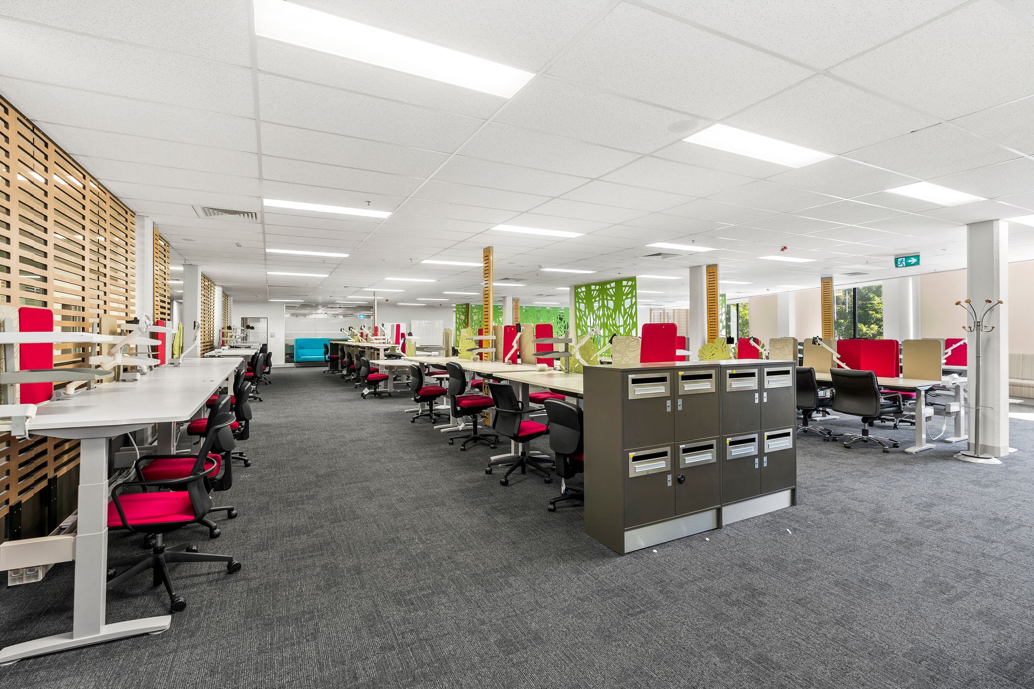 This is a modern open-plan office space with shared workstations, ergonomic chairs, and a well-lit environment. The red and black chairs contrast with the neutral decor, and there are partitions with green decorative elements. It looks like a collaborative workspace designed for productivity.
