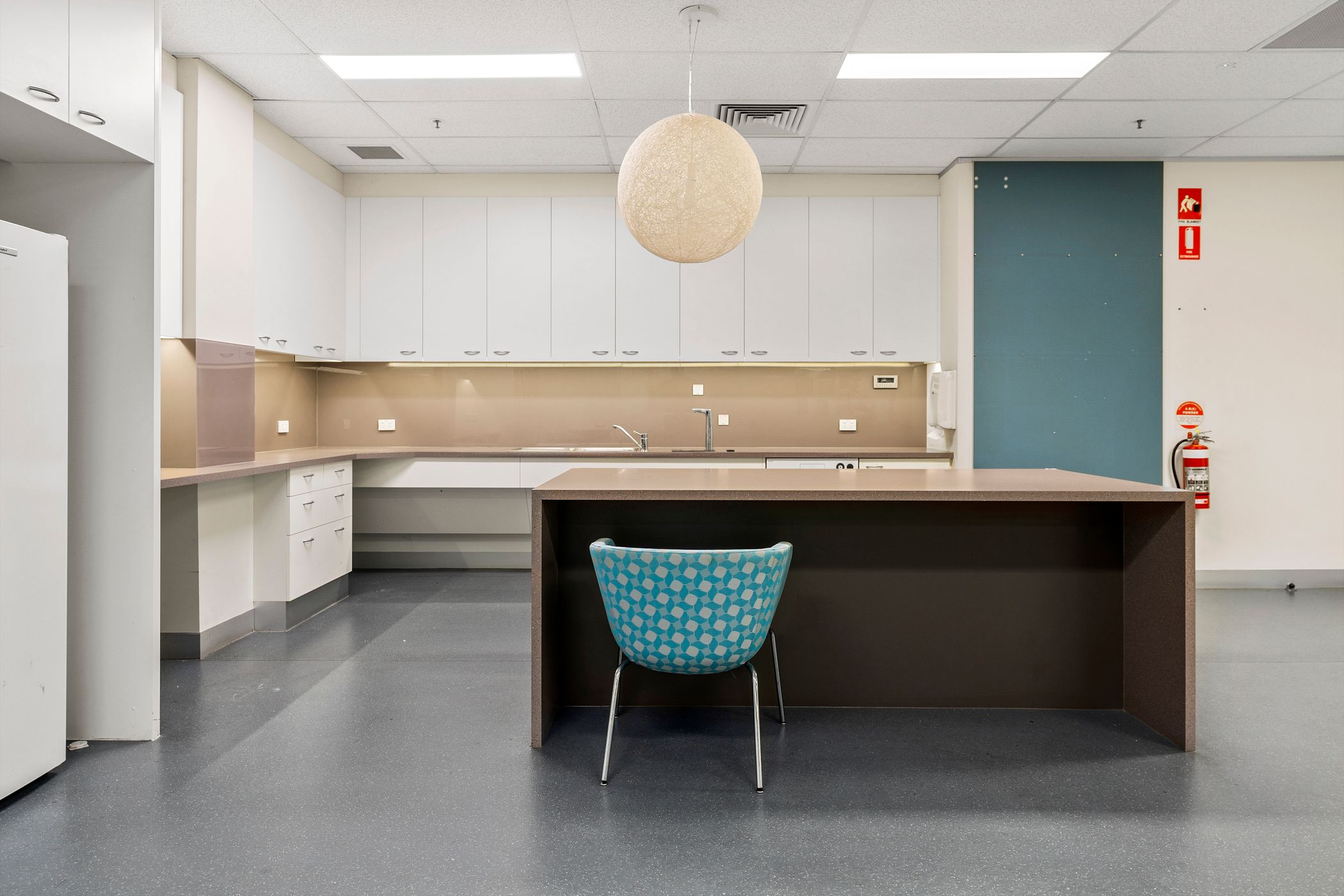 Small kitchen space, white cupboards surround the room with a sink and taps, in the centre of the room is an island bench with a blue chair pulled up to it, above is a large round light, Hamilton Group Geelong