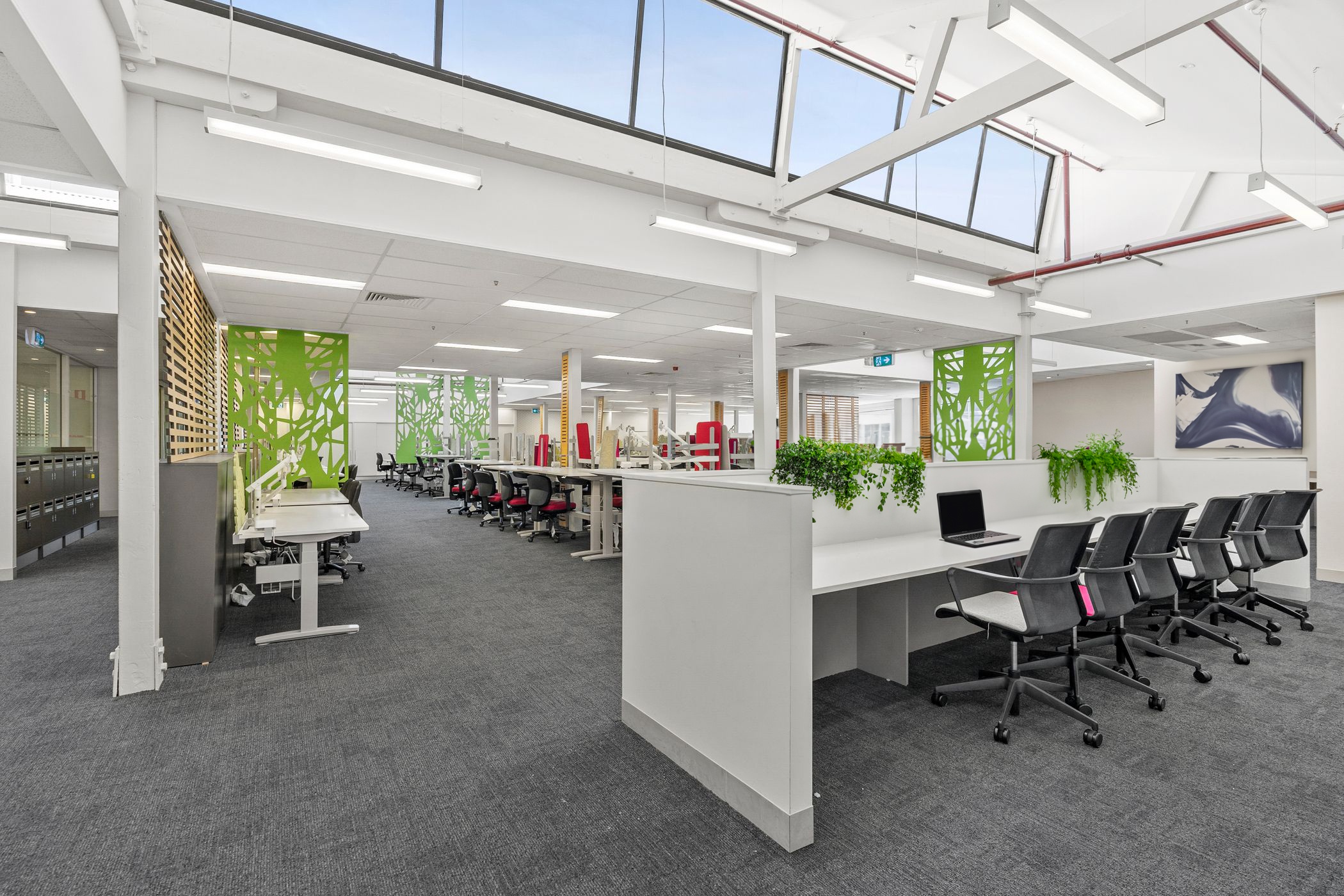 A modern open-plan office space with multiple rows of workstations, ergonomic black and red chairs, and white desks. The office features green decorative partitions, skylights allowing natural light, and overhead fluorescent lighting. There are lockers for storage, potted plants adding greenery, and a mix of collaborative and individual workspaces. The floor is carpeted in dark grey, and the walls are adorned with wooden paneling and artwork