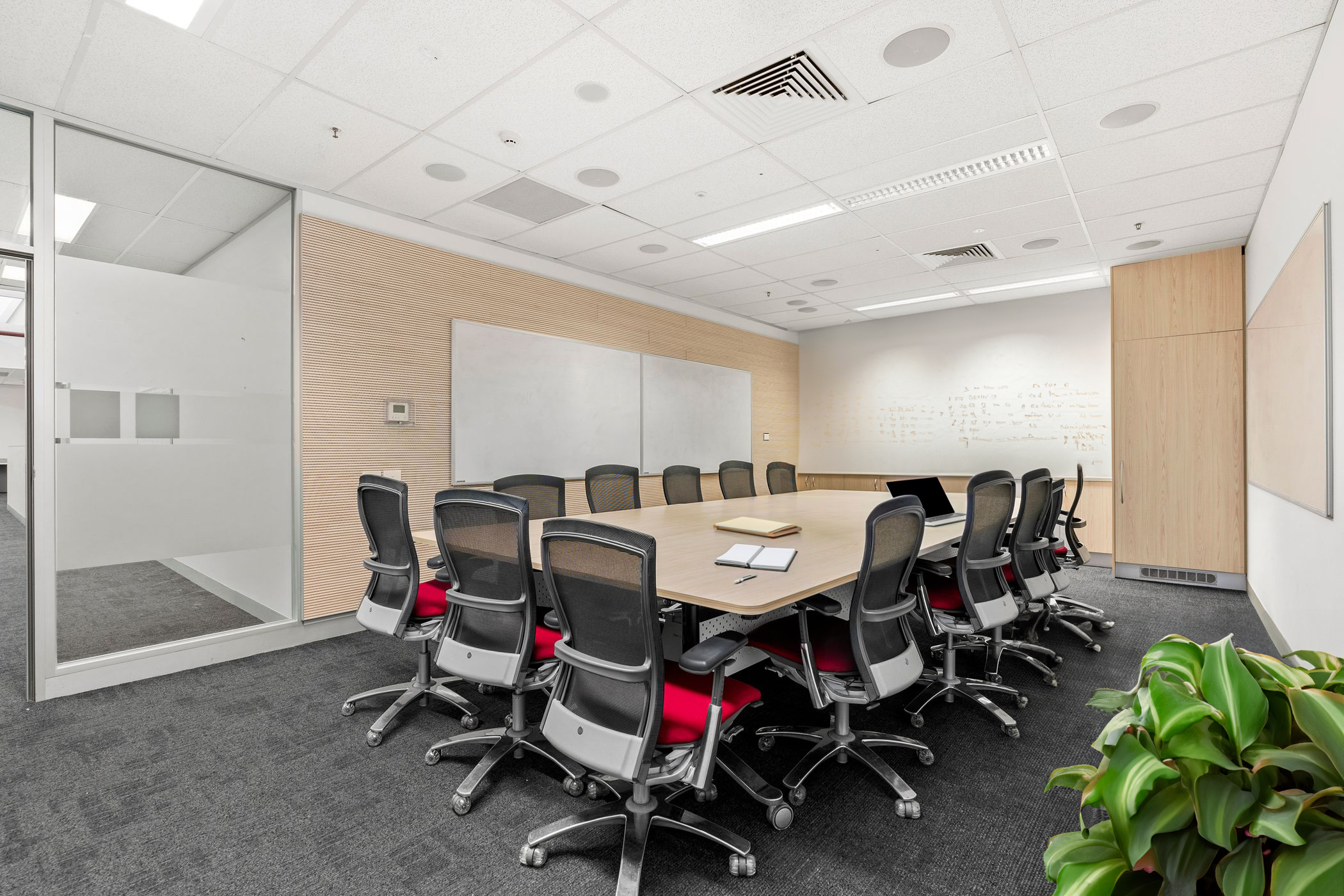 A modern open-plan office space with multiple rows of workstations, ergonomic black and red chairs, and white desks. The office features green decorative partitions, skylights allowing natural light, and overhead fluorescent lighting. There are lockers for storage, potted plants adding greenery, and a mix of collaborative and individual workspaces. The floor is carpeted in dark grey, and the walls are adorned with wooden paneling and artwork