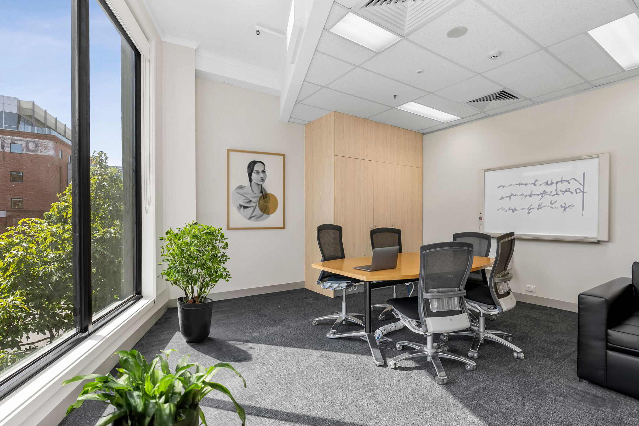 A modern, well-lit meeting room with a large window offering a view of trees and a brick building. The space features a round wooden table surrounded by ergonomic office chairs, a whiteboard with abstract writing, and framed artwork of a stylised portrait. The room is decorated with potted plants and has a neutral colour palette with wooden and black accents, creating a professional yet inviting atmosphere.