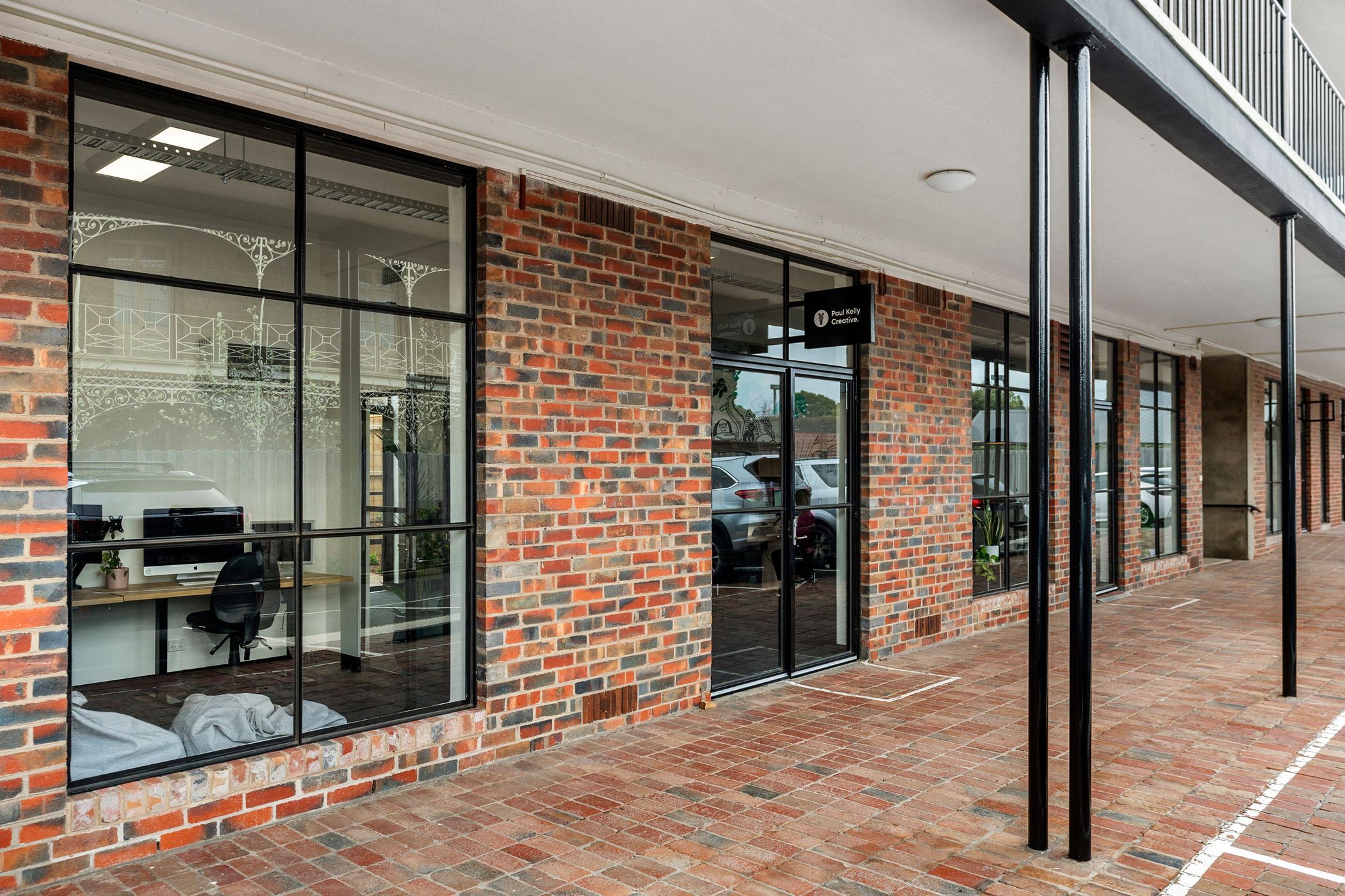 Red brick facade building with many multi colour bricks filled in between, black tbar windows and doors line the facade, inside the building is a small office space with a lone chair and computer, a second story above shows that there is a balcony above, Hamilton Property Group