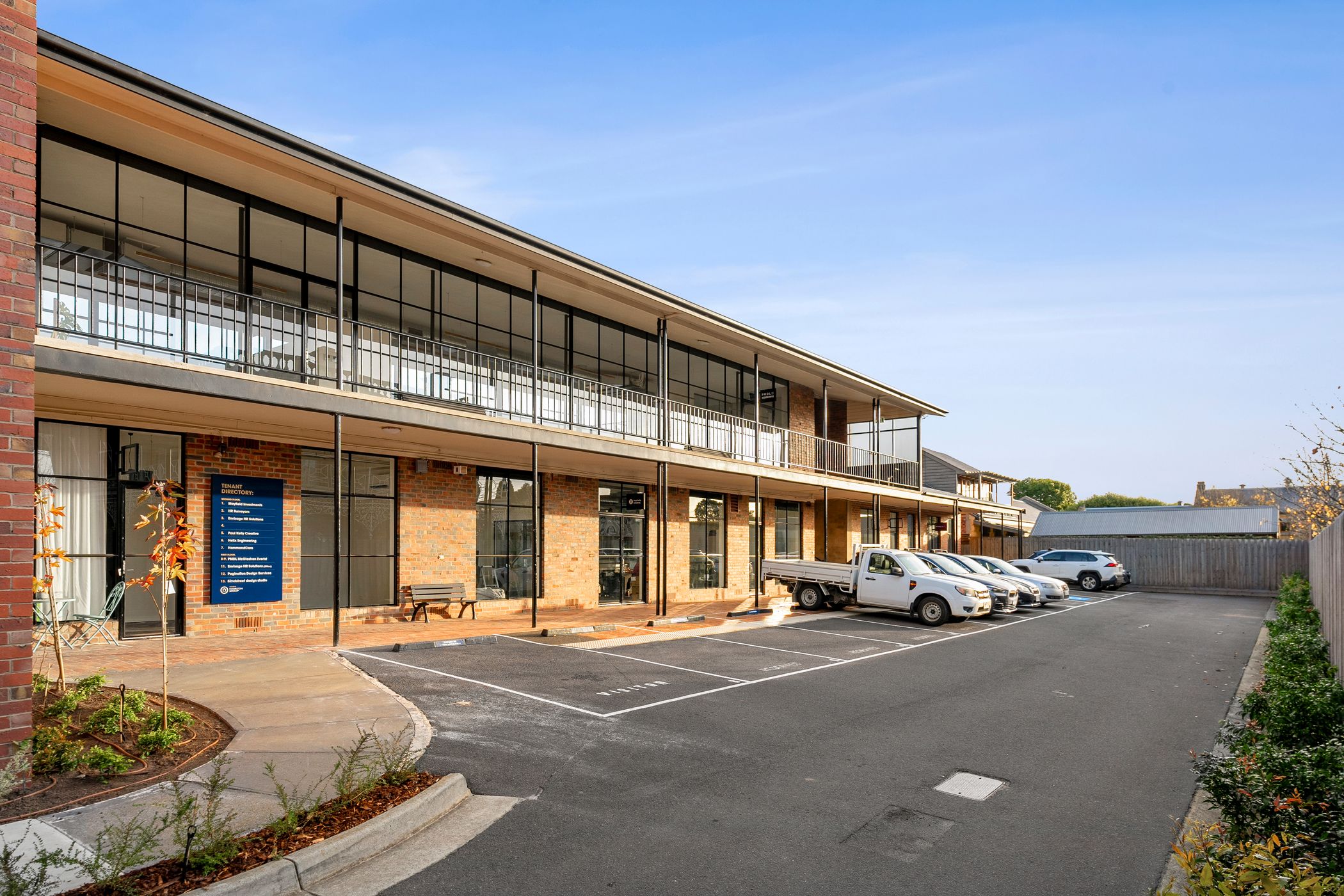 Car park view of a large double story building, and orange brick is used for the facade, tbar windows and doors and spread throughout, the car park has six cars parked within it, and theres clear blue skies over-head, Hamilton Property Specialists
