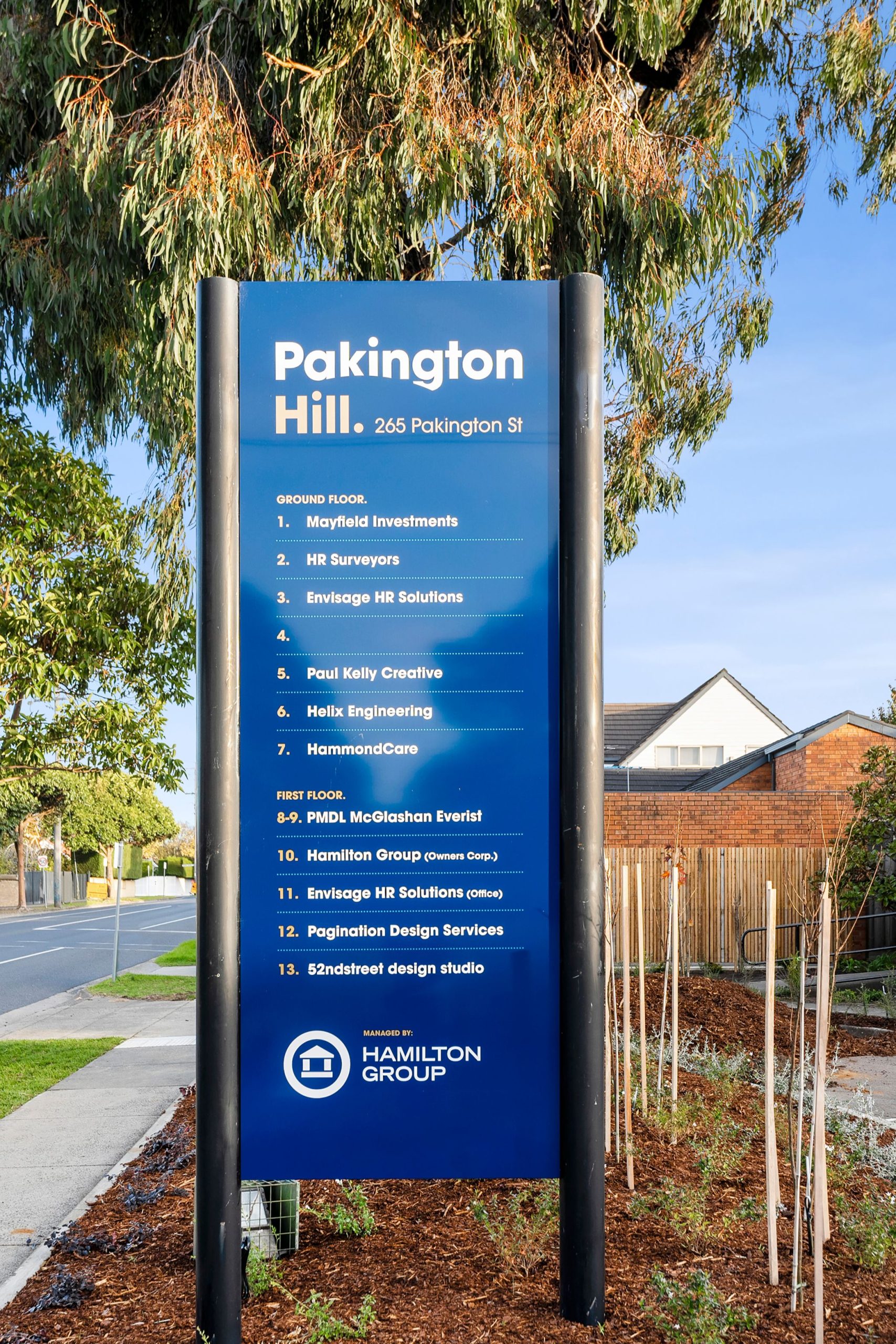 Tall directory sign located at 265 Pakington Street, the sign is blue in colour with two black posts on either side holding it up, the sign has directions to two floors of a building, on the top it states "Pakington Hill" and at the bottom it has "Hamilton Group" in the background it has a large tree, a small house and clear blue skies above, Hamilton group Geelong