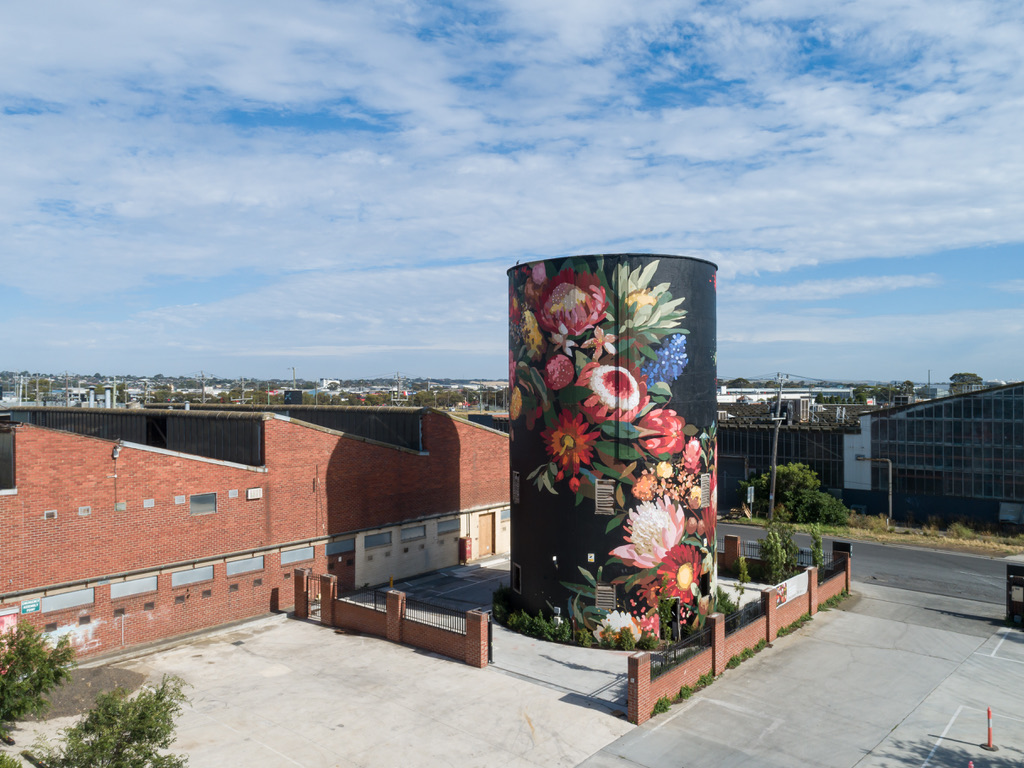 A vibrant floral mural covers a tall, cylindrical tower in an Federal Mills, North Geelong, surrounded by brick buildings and paved surfaces under a partly cloudy sky, blending urban structure with natural beauty. Hamilton Group