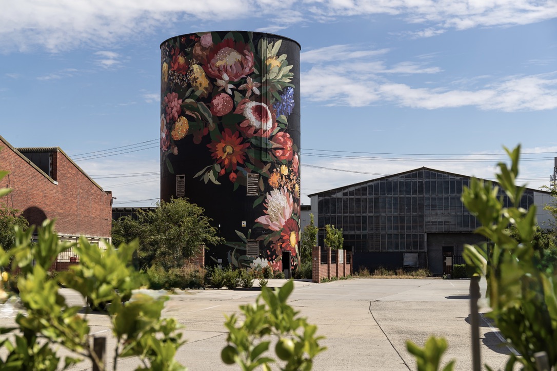 A large cylindrical silo painted with a vibrant mural of native Australian flowers, set within an industrial precinct at Federal Mills in North Geelong. The background features a blue sky with scattered clouds and surrounding heritage brick and glass factory buildings. Green foliage frames the foreground.